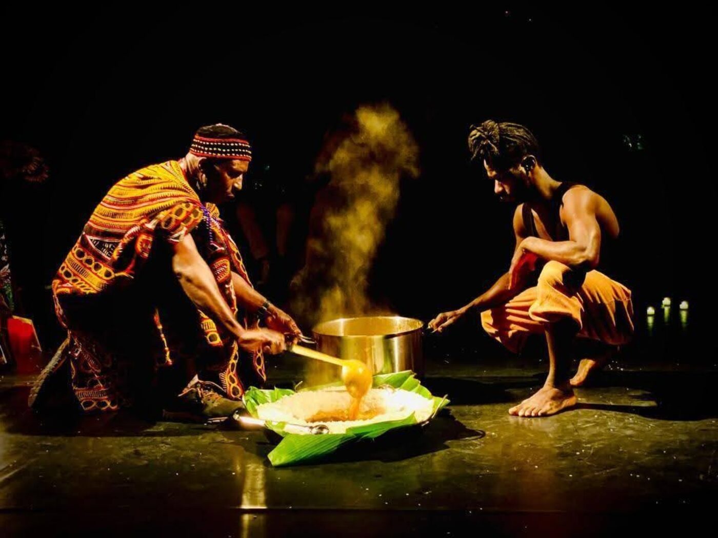 Two men in traditional attire prepare food together onstage, with steam rising from a pot and a dish served on banana leaves in front of them, under dramatic lighting.
