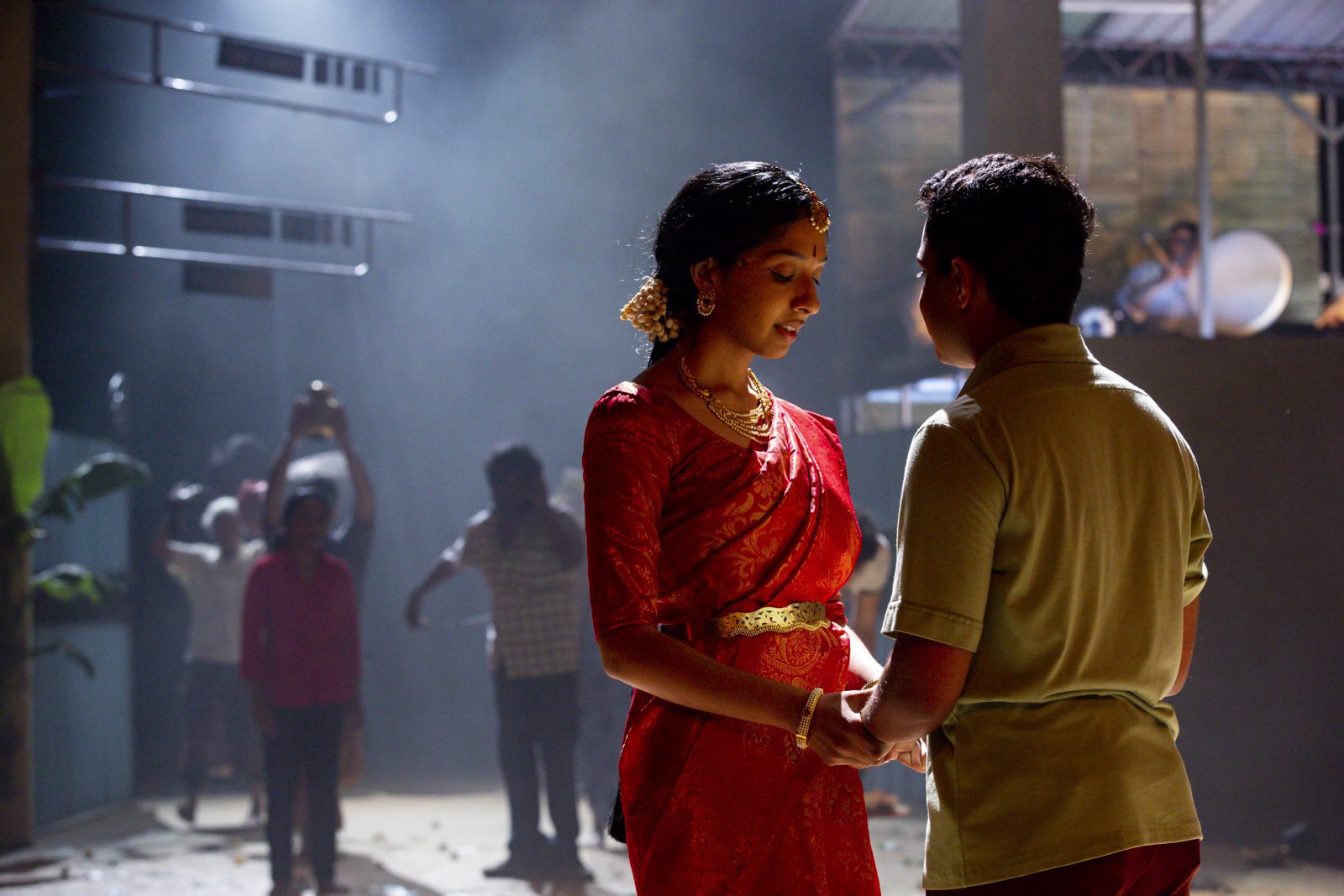 A woman in a red saree and a man in a green shirt hold hands, facing each other, while people watch in the background amid dramatic lighting and some scattered debris.