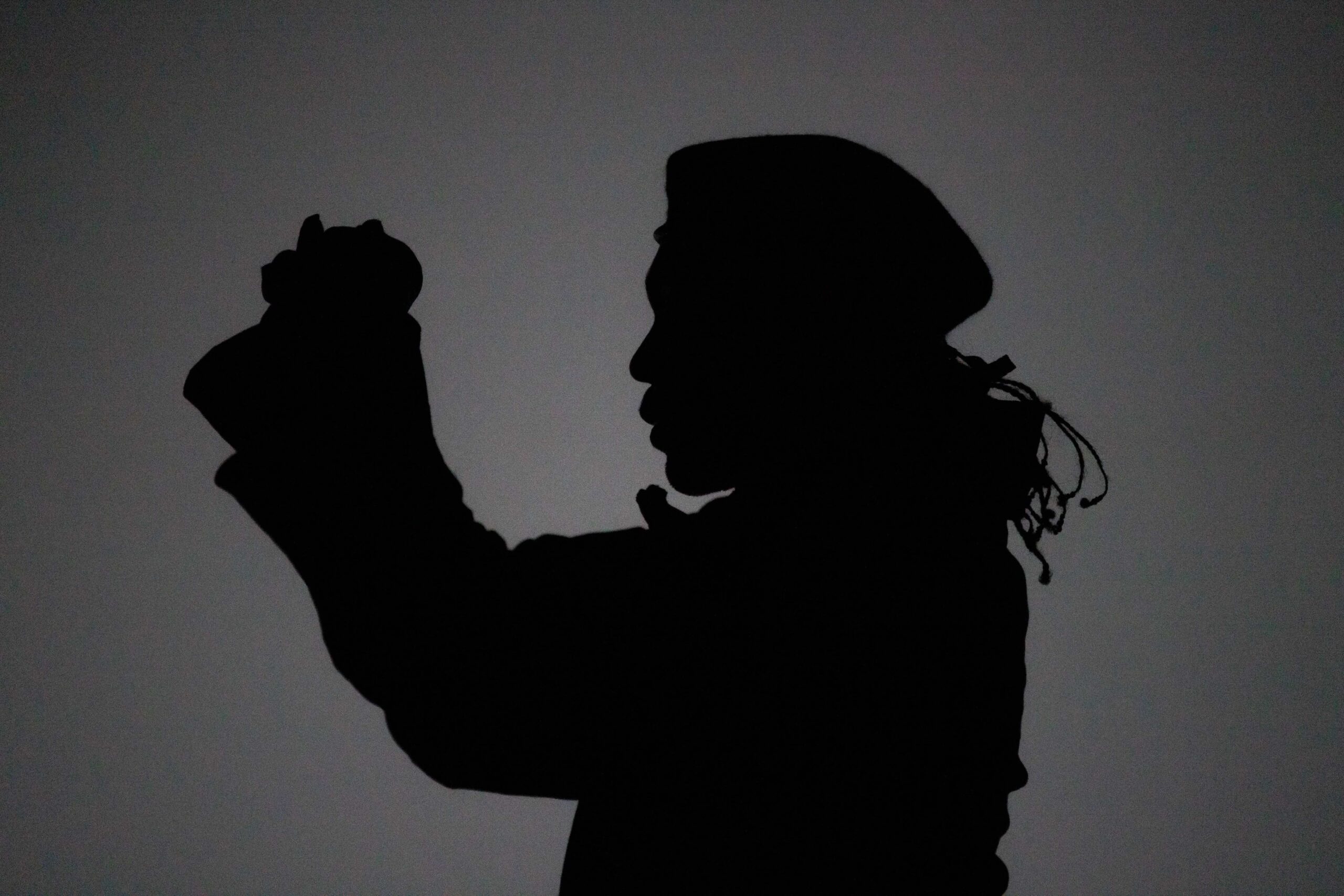 Silhouette of a person wearing a hat or head covering, holding their hands together and raised, set against a plain light background.