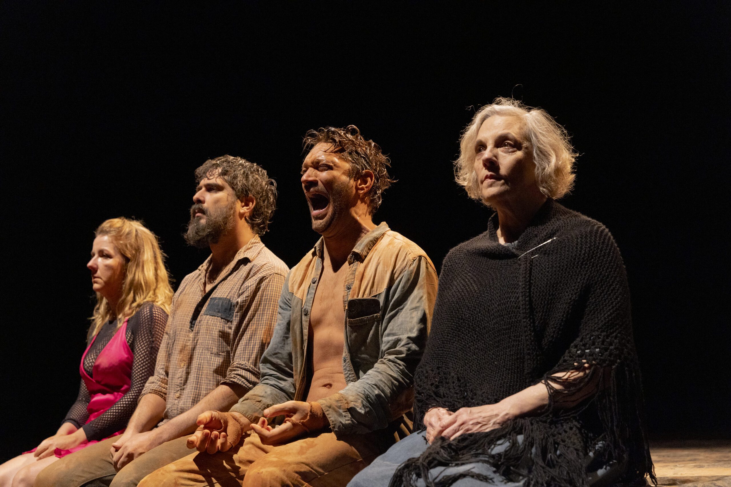 Four people sit in a row onstage under dramatic lighting; the two men in the middle appear distressed, with one yelling, while the women on each end look somber and contemplative.