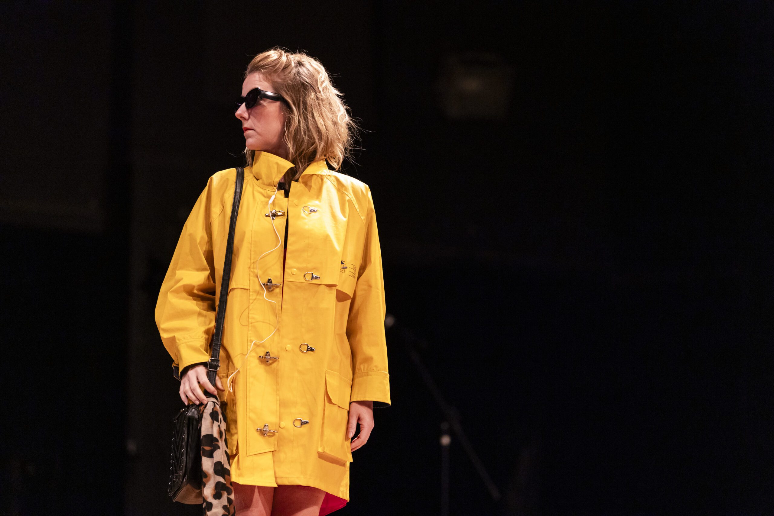 A person wearing a bright yellow coat and sunglasses stands on stage holding a leopard print bag, looking to the side against a dark background.