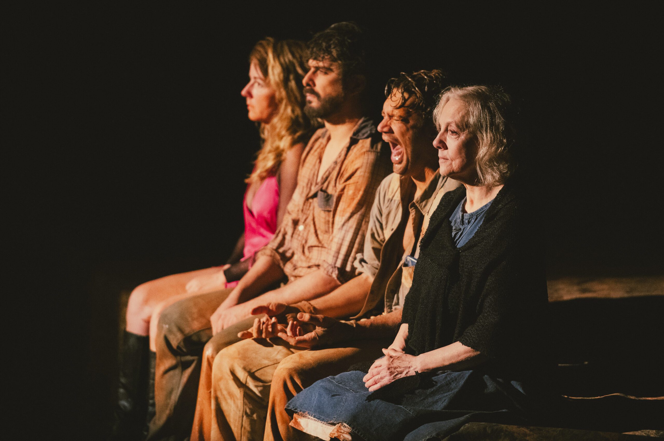 Four people sit in a row onstage, looking forward. Three have neutral expressions, while the third person from the left appears to be yelling or singing. The lighting is dramatic, highlighting their faces.