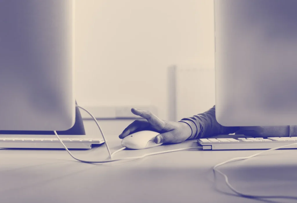 A person’s hand uses a computer mouse between two monitors on a desk, with visible cables and a keyboard, in a softly lit, modern workspace.