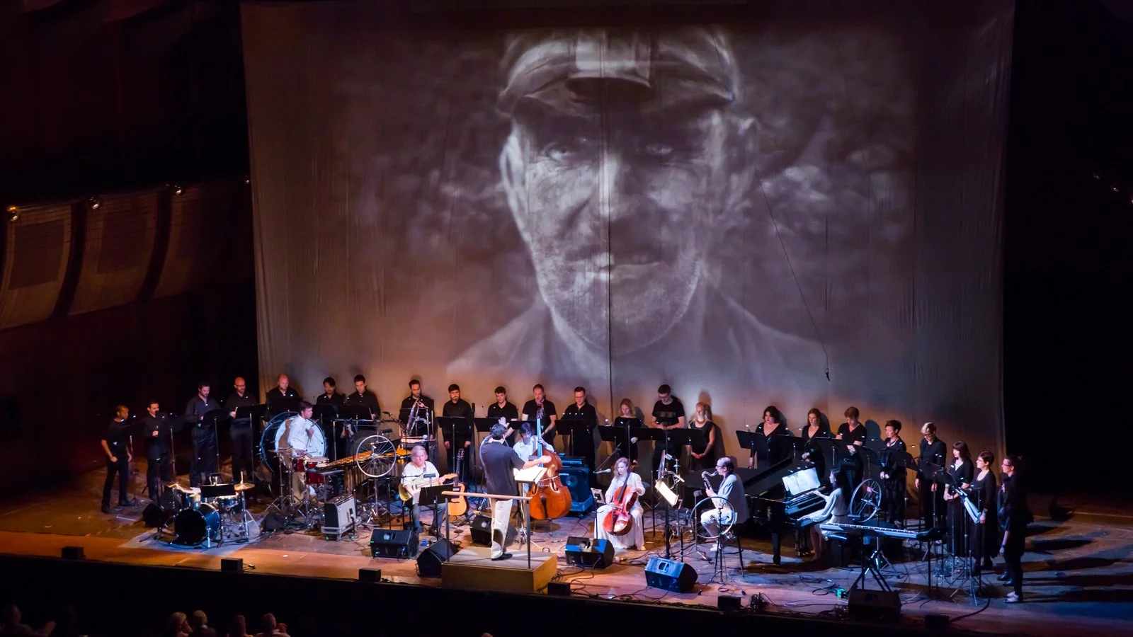 A large orchestra and choir perform on stage with a big black-and-white image of a man’s face projected behind them, creating a dramatic atmosphere.