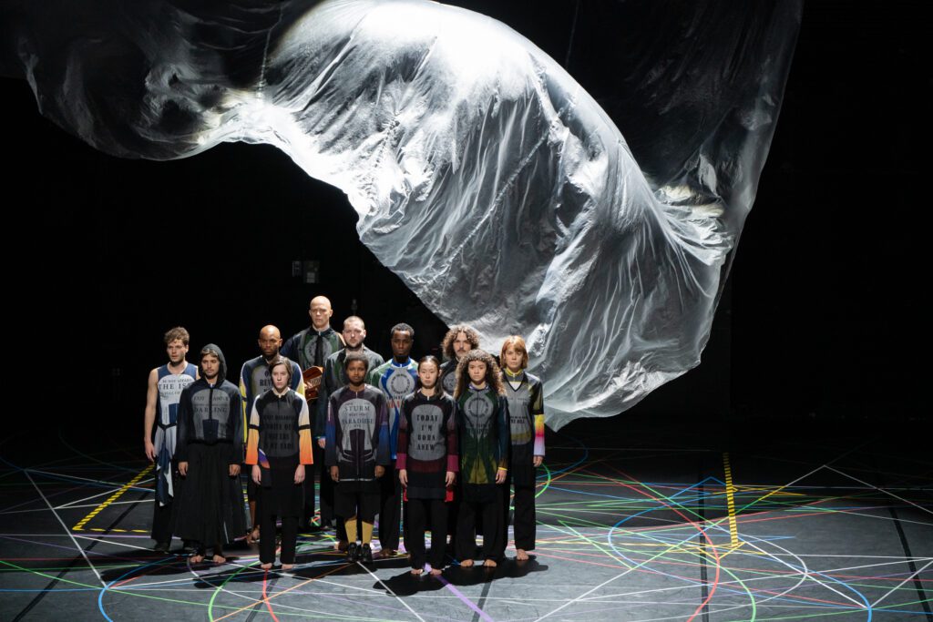 A group of people in matching outfits stand in formation on a dark stage with colorful lines, beneath a large, suspended, translucent sheet.
