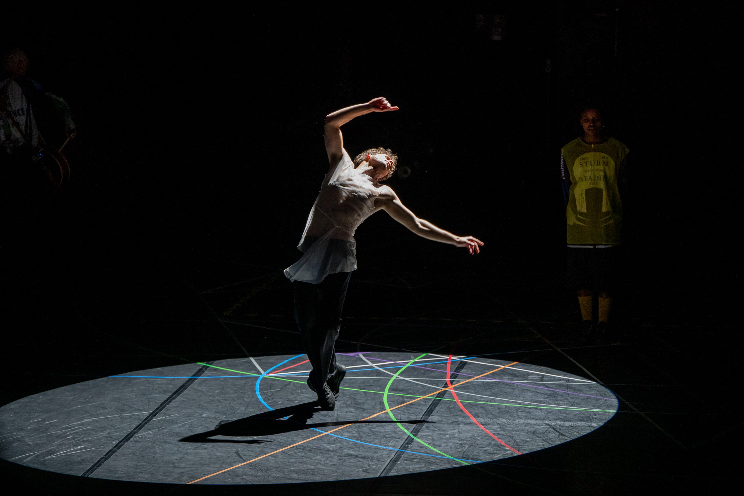 A dancer in a sheer white top performs under a bright spotlight on a dark stage with colorful lines on the floor, while another person in a yellow shirt watches from the shadows.