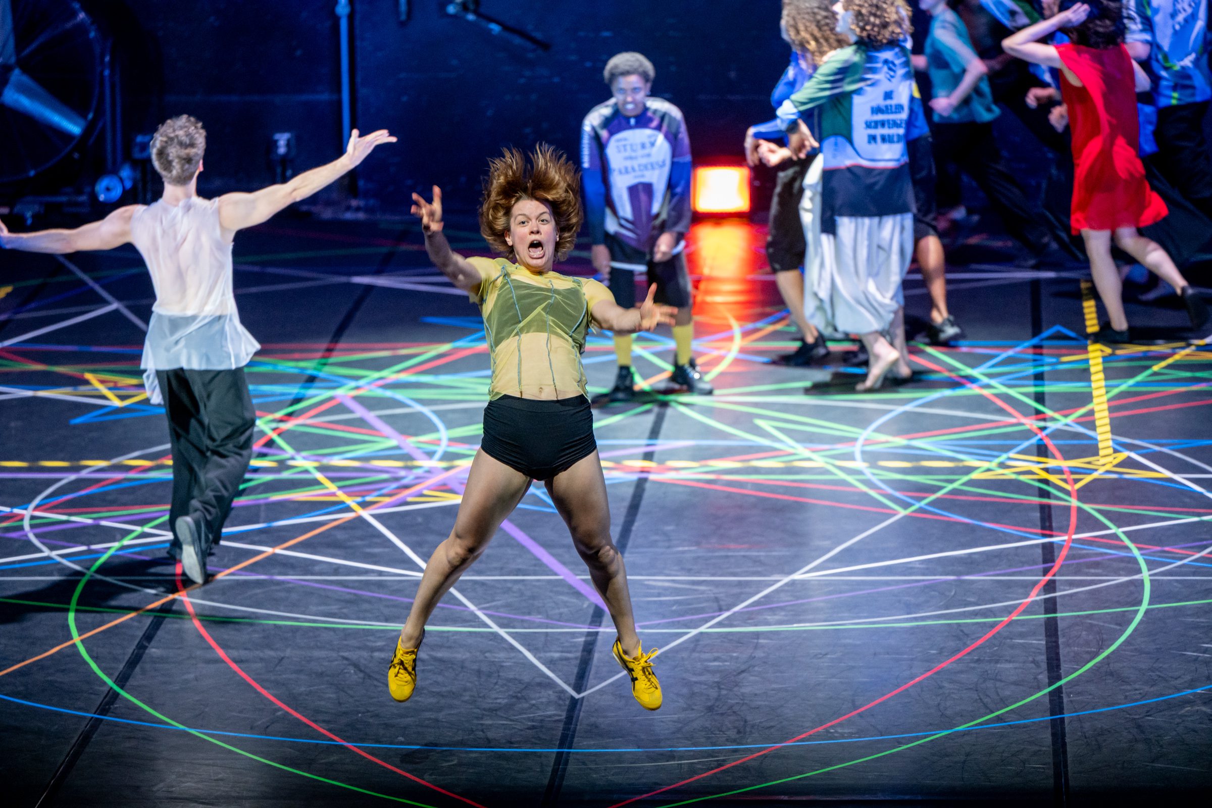 A person jumps energetically center stage, surrounded by colorful lines on the floor. Other performers in varied outfits move in the background. The scene appears lively and dynamic.
