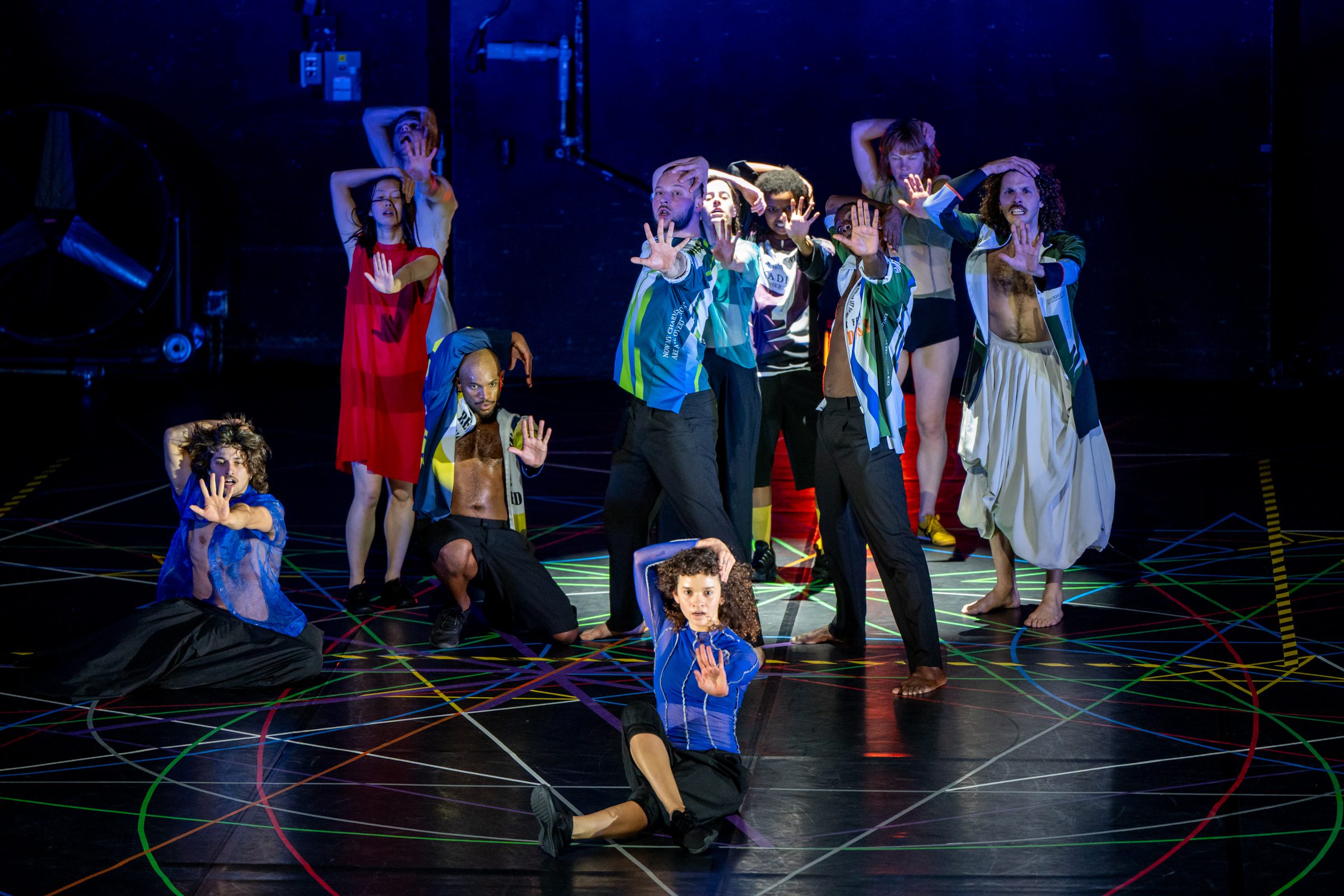 A group of dancers strike dramatic poses on a stage, with colorful lines on the floor and expressive arm movements, set against a dark background.