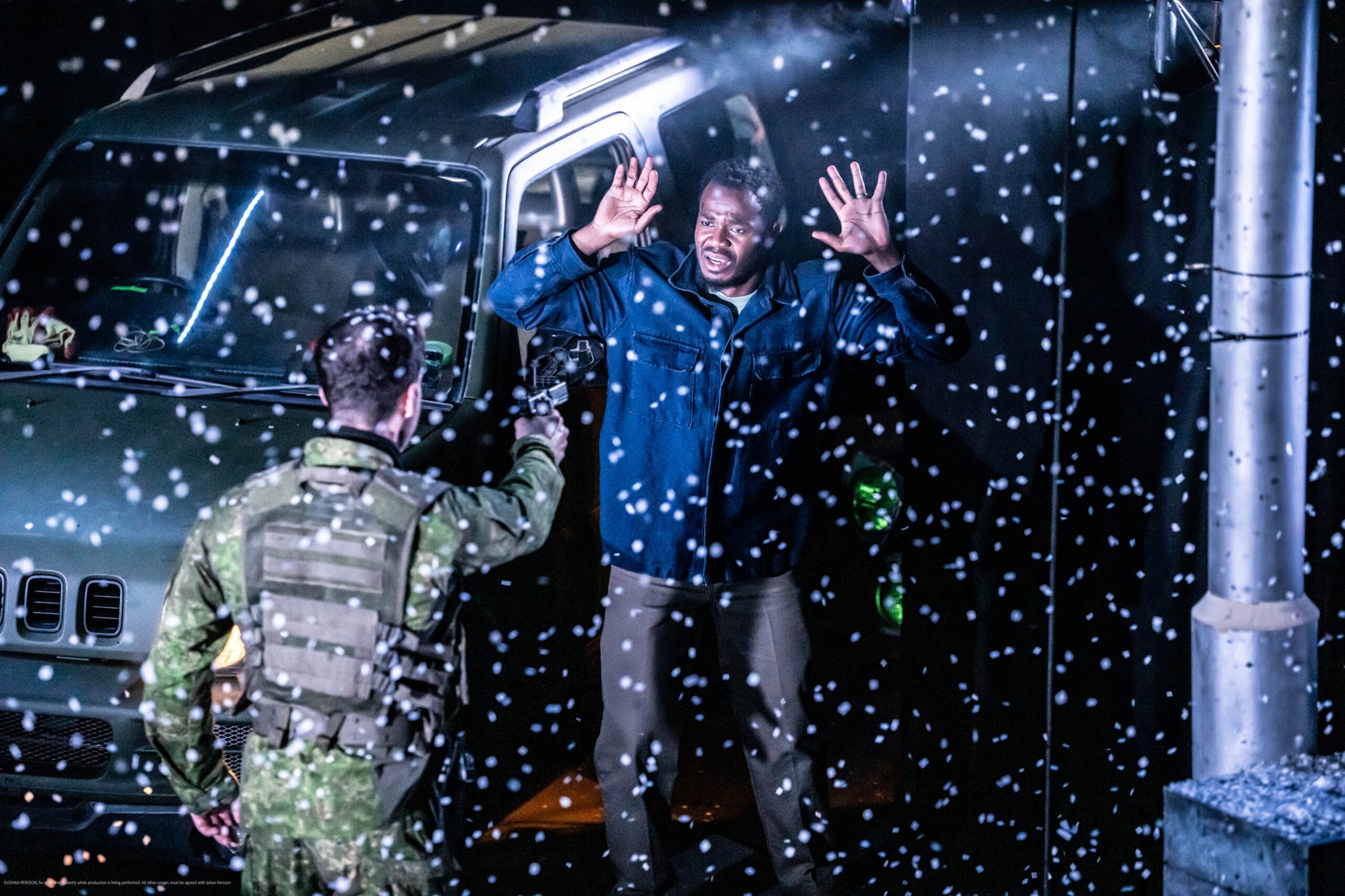 A soldier points a gun at a man with raised hands beside a green vehicle, while white particles or raindrops are visible in the air around them at night.