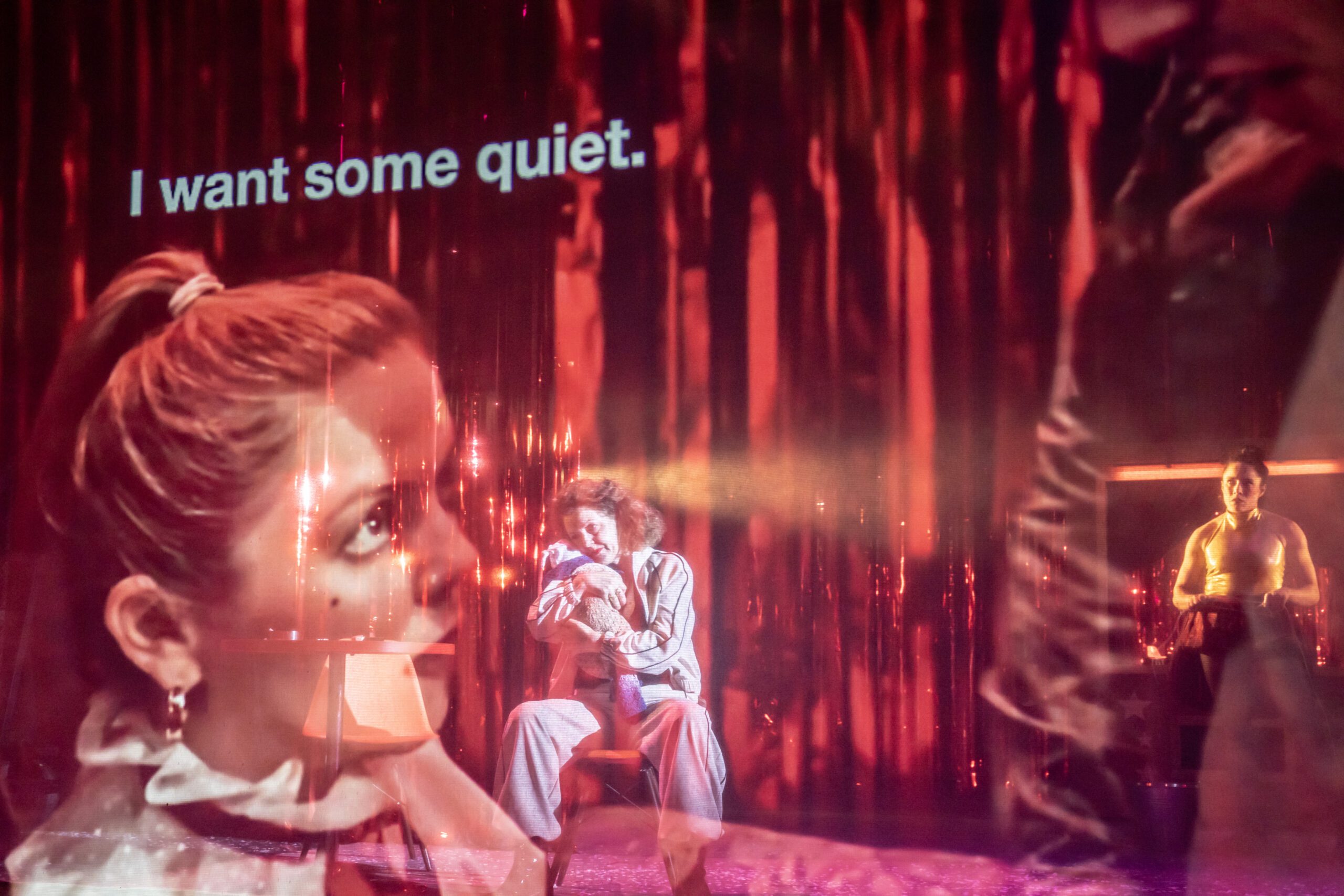 A stage scene bathed in red with layered images of performers. Large on-screen text reads, “I want some quiet.” One performer sits in the center while others stand and gesture in dramatic theatrical lighting.