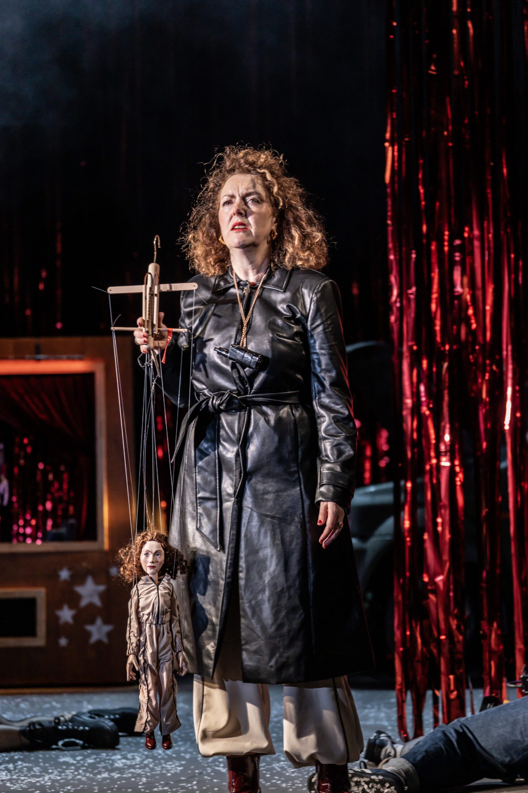 A woman in a black leather coat holds a marionette doll. She stands on stage with red metallic streamers in the background and another person lying on the floor behind her.