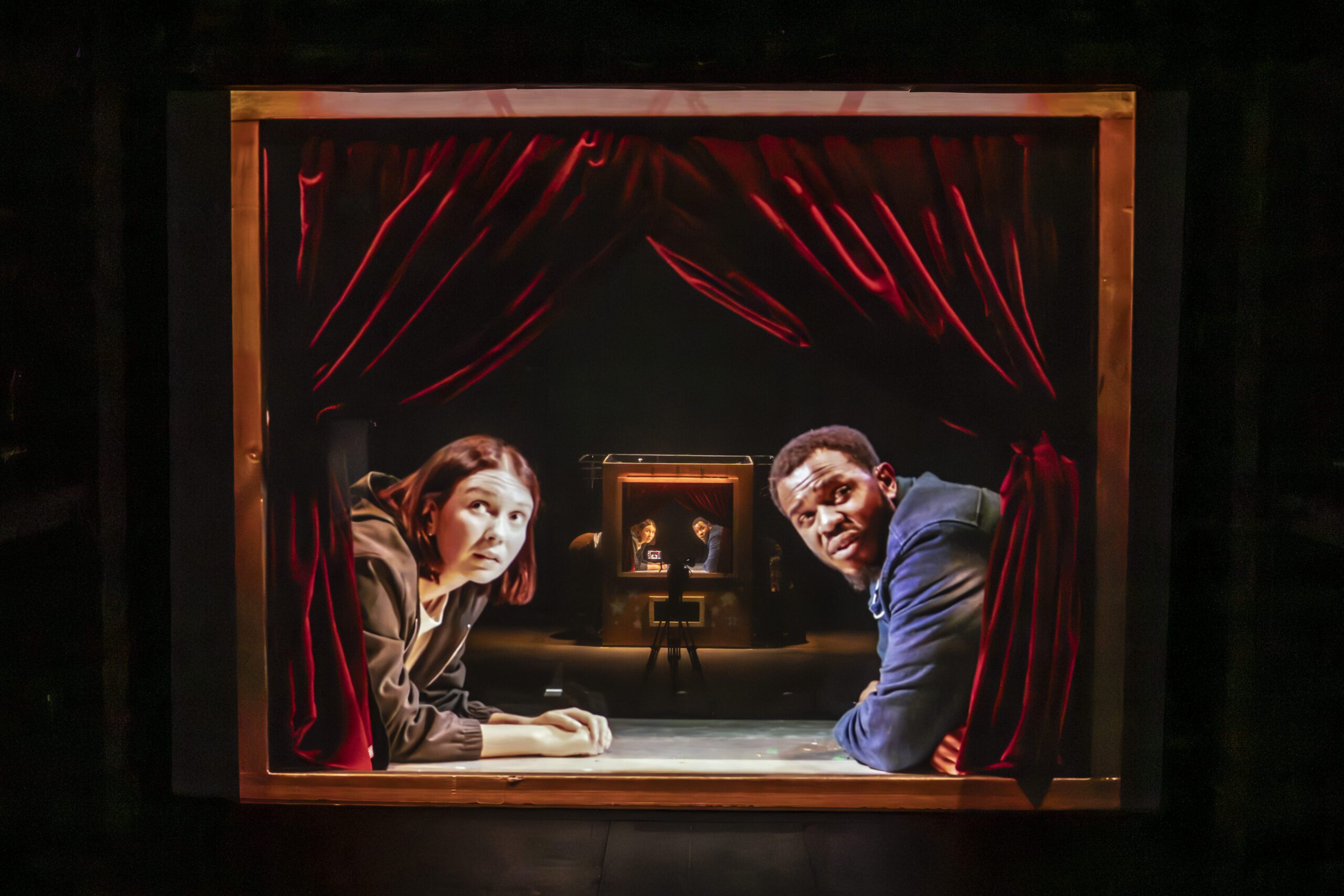Two people lean on a stage frame with red curtains, looking toward the camera. In the background, a smaller version of the same scene repeats, creating a tunnel-like effect.