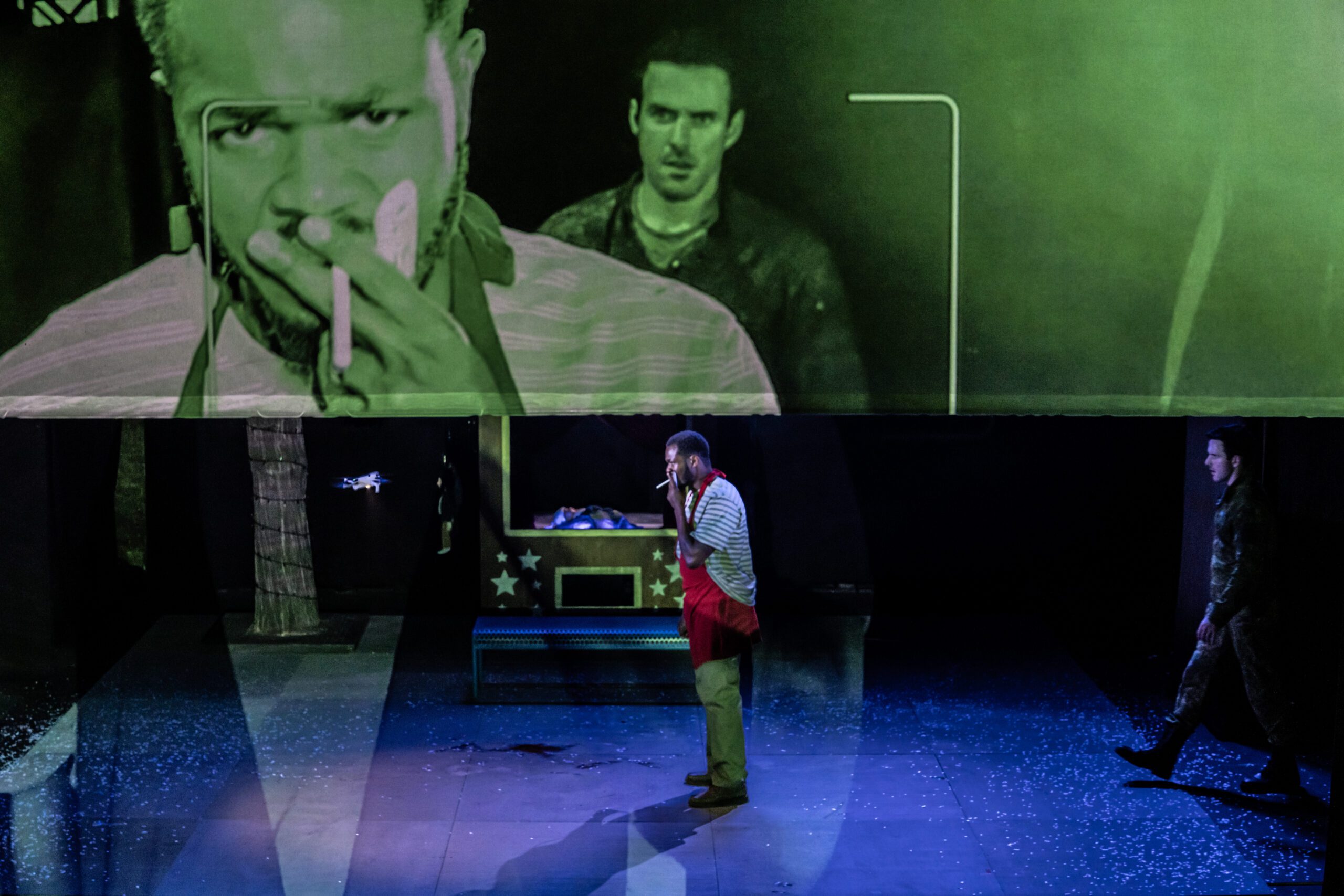 A man smokes onstage under a large screen showing his close-up. Another man stands in the background, watching him. The stage is dimly lit, with scattered props and a blue-lit floor.