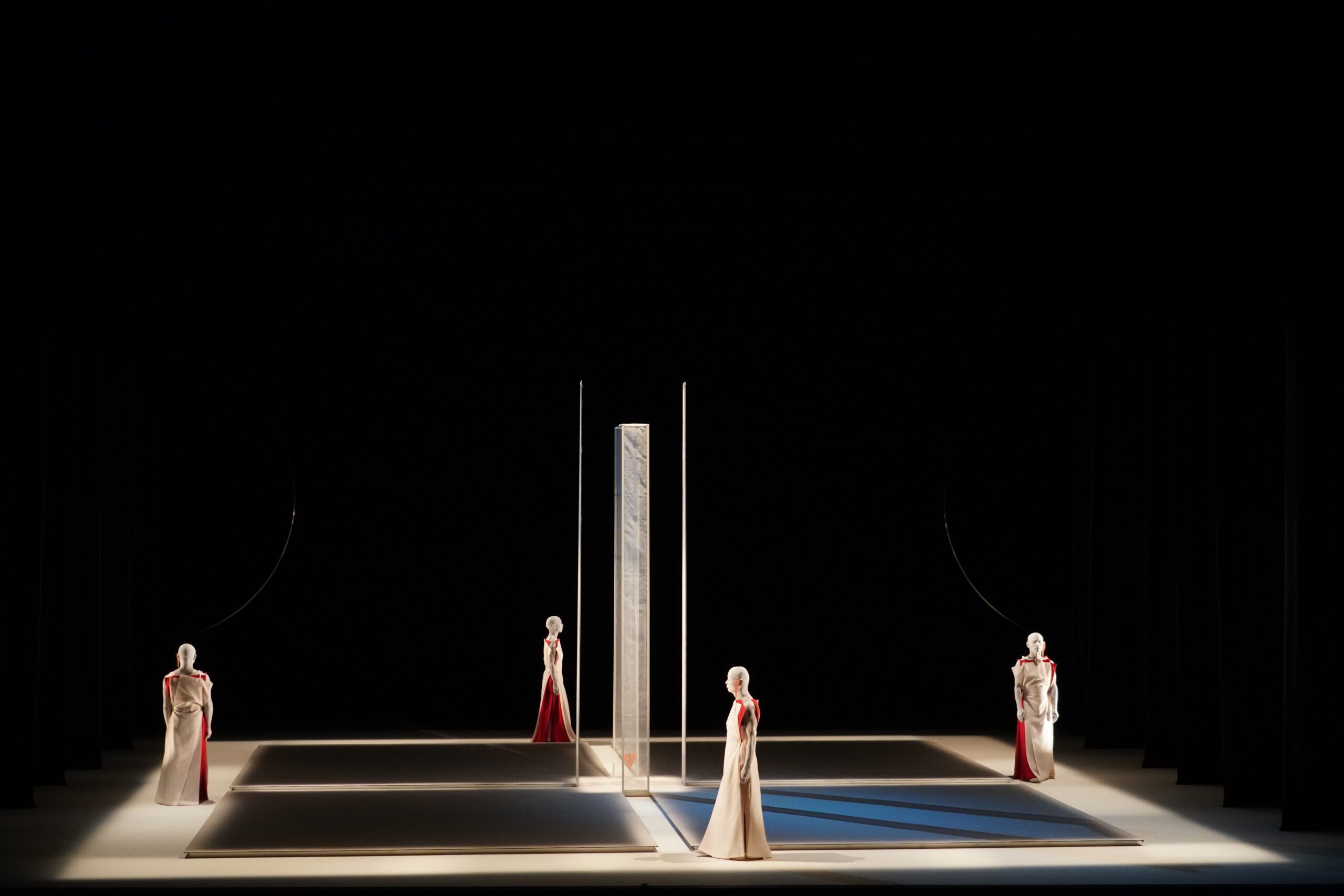 Four figures in long white and red gowns stand on a minimal, modern stage with a tall, mirrored panel at the center, dramatic lighting, and strong shadows against a dark background.