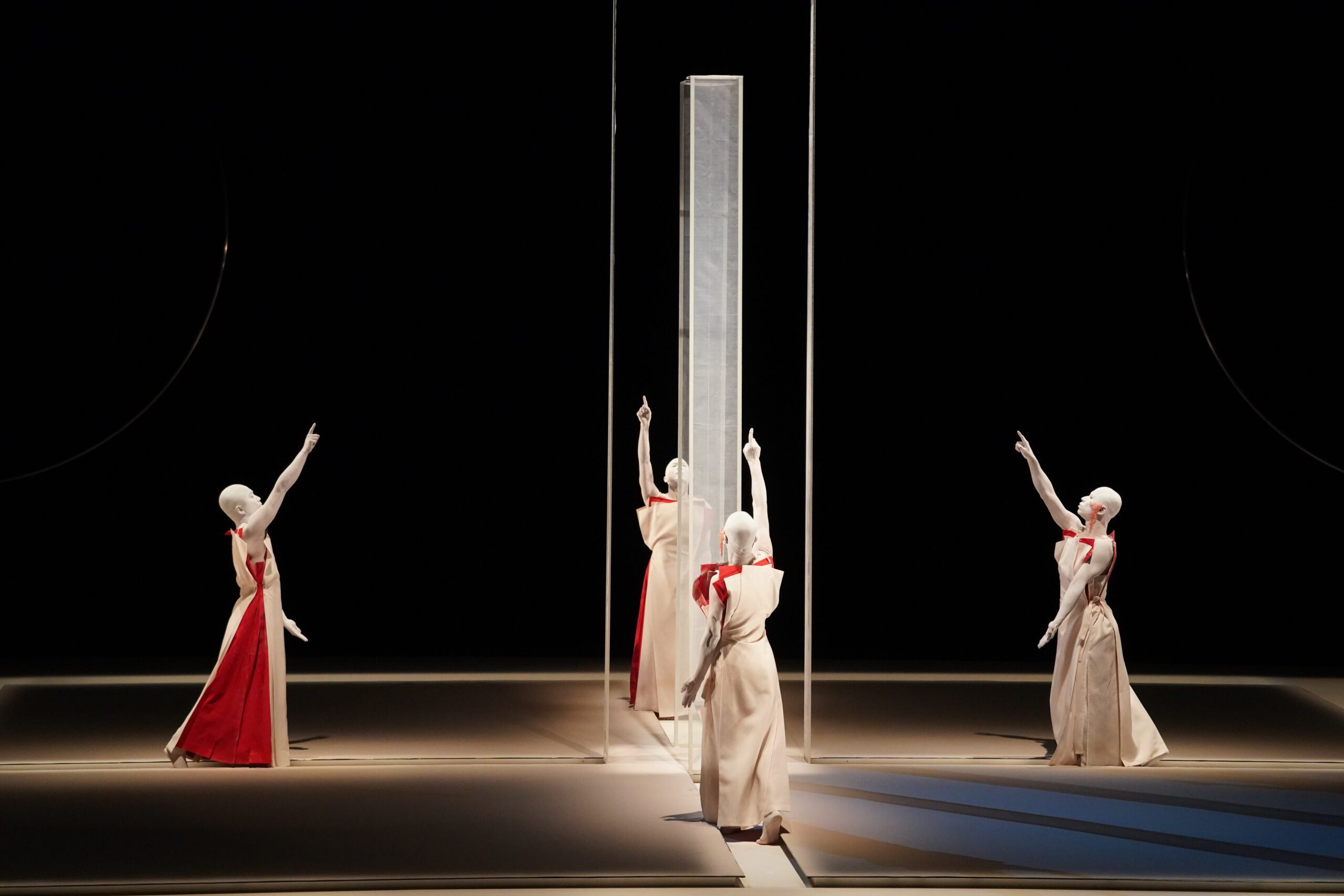 Four dancers in flowing white and red costumes stand around a tall transparent pillar on stage, each extending one arm upward, with dramatic lighting emphasizing their poses.