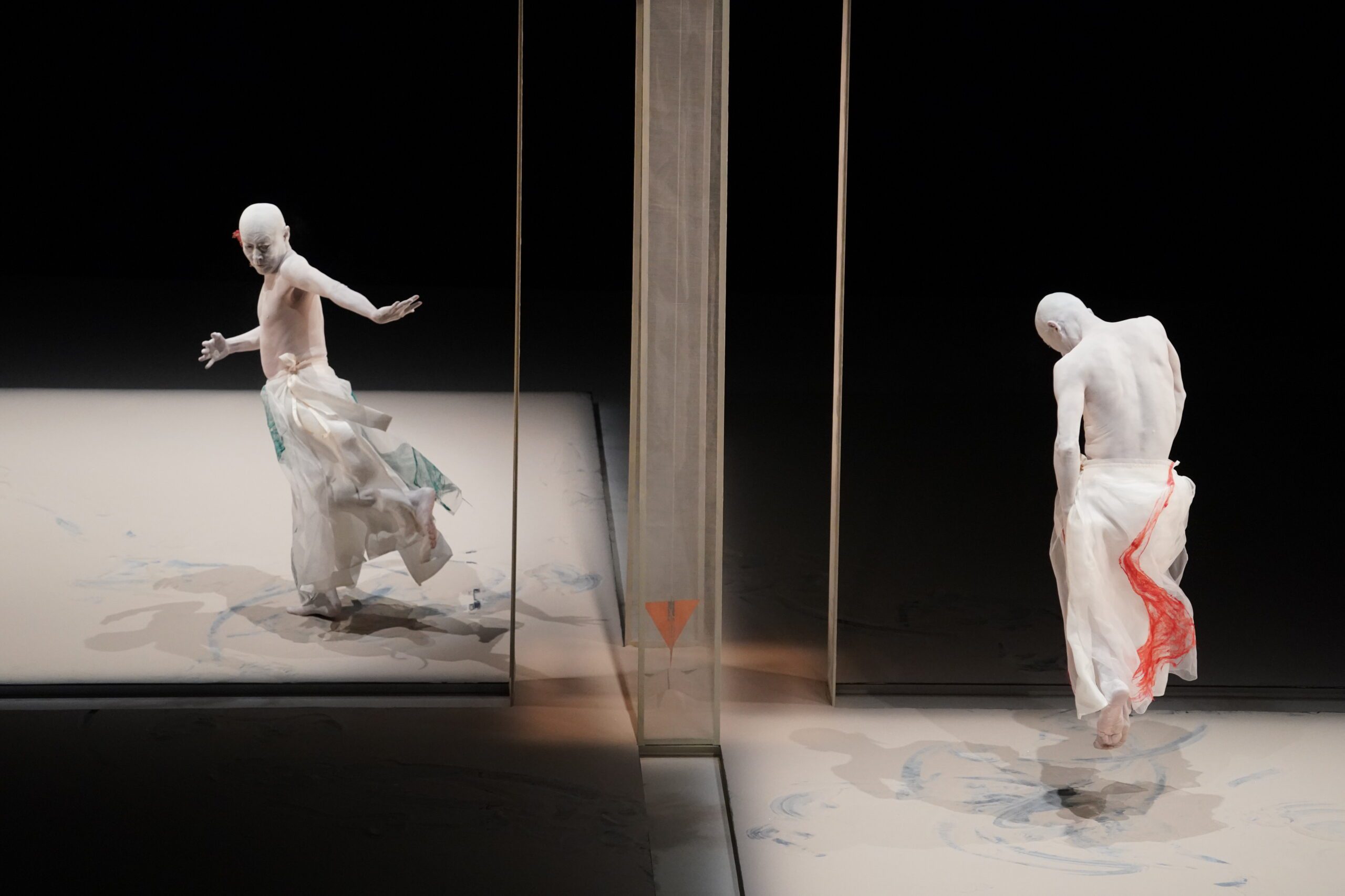 Two performers with white-painted skin and flowing skirts dance on a pale stage, separated by a transparent panel. One appears mid-motion while the other stands still, both reflected in the surface.