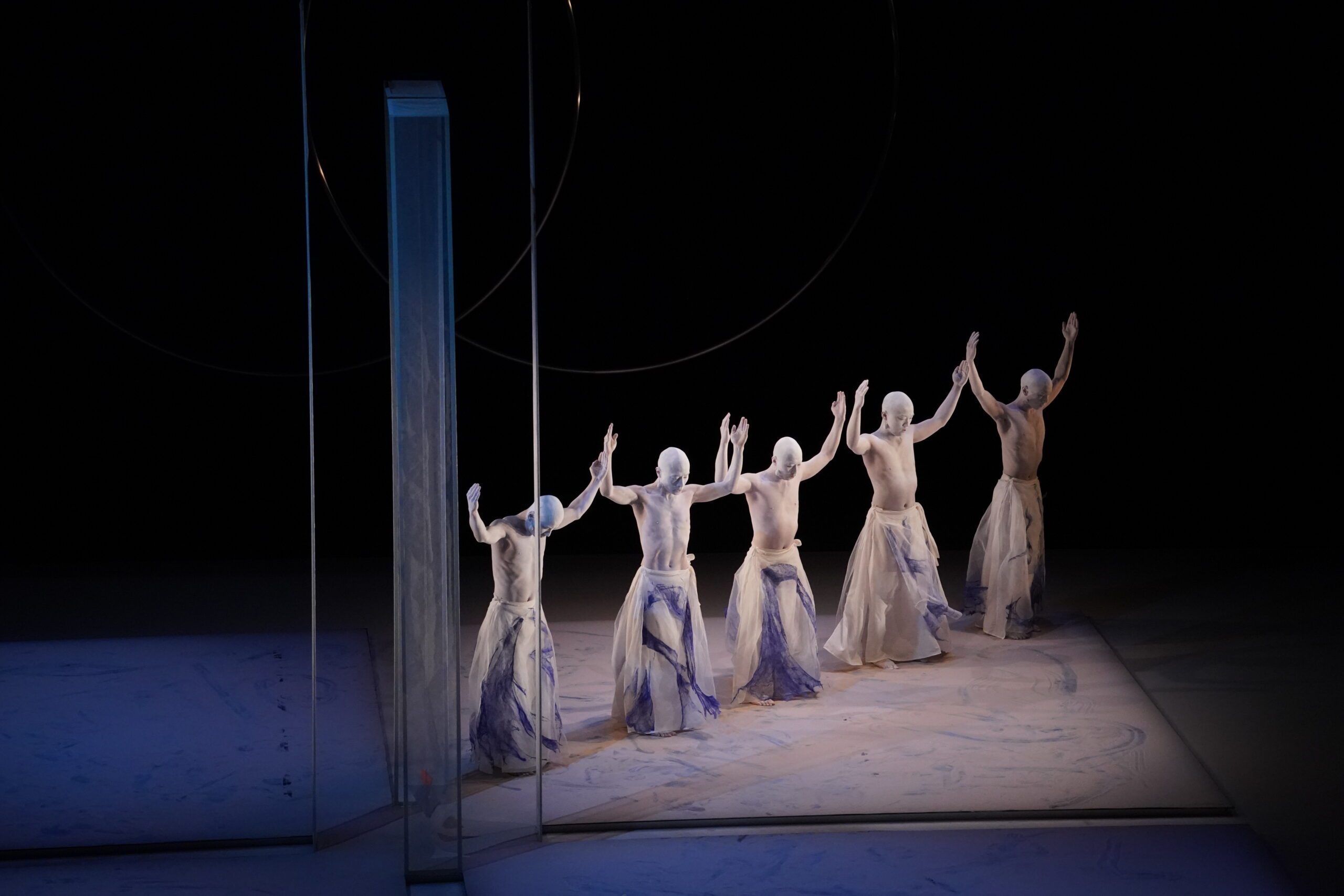 Five dancers wearing white costumes and head coverings pose with arms raised, standing in a row on a dimly lit stage with a vertical reflective panel beside them.