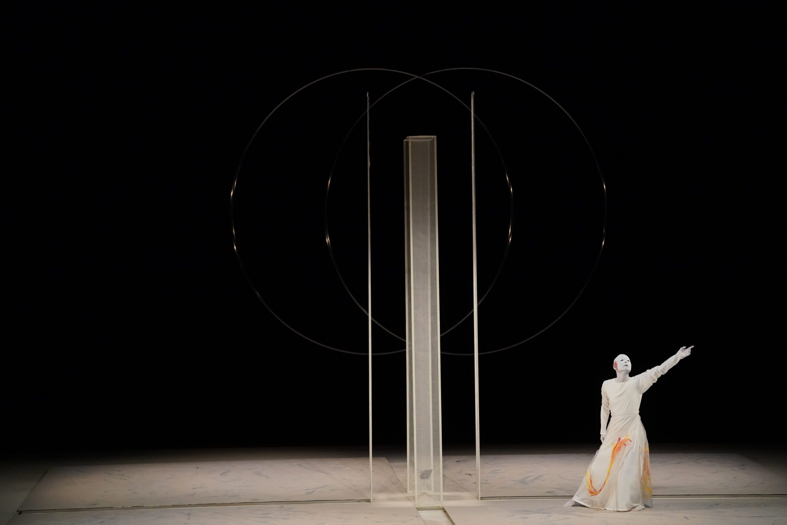 A performer in white costume with orange accents poses on stage beside minimalist set pieces: a tall rectangular frame and two intersecting circles, against a dark background.