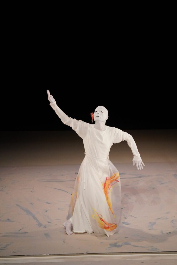 A performer in a white mask and flowing white costume with orange accents poses dramatically on stage, one arm raised and the other bent, against a neutral background.