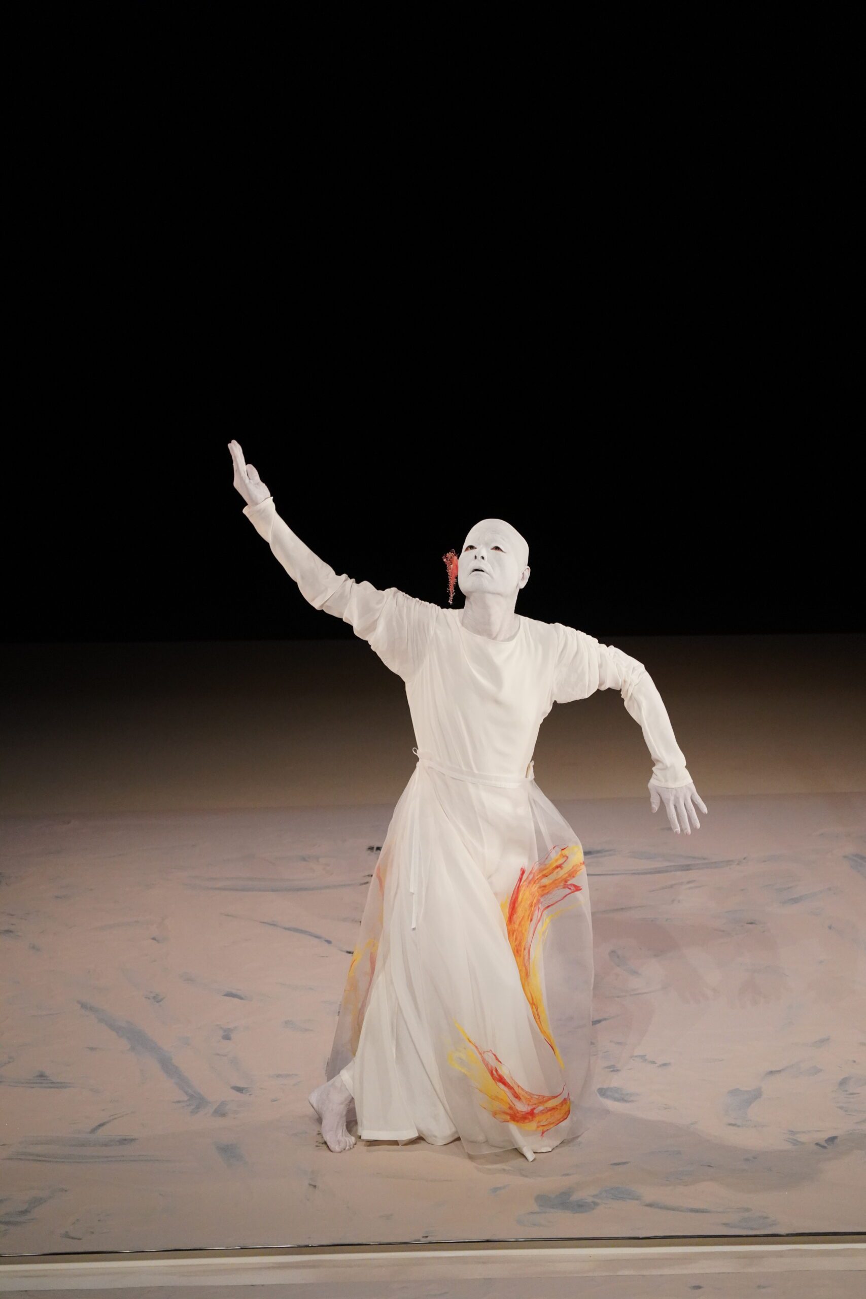A performer in a white mask and flowing white costume with orange accents poses dramatically on stage, one arm raised and the other bent, against a neutral background.
