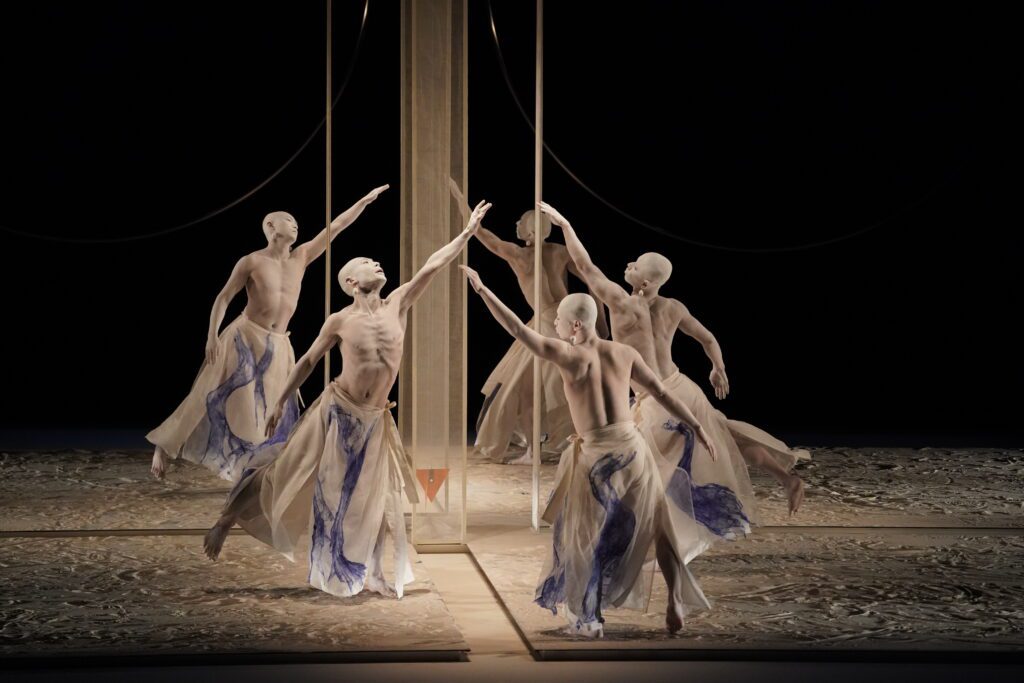 Five dancers with bald heads and flowing cream and blue skirts perform a synchronized, expressive dance around a vertical structure on a textured stage, reaching out with extended arms in dramatic poses under soft lighting.