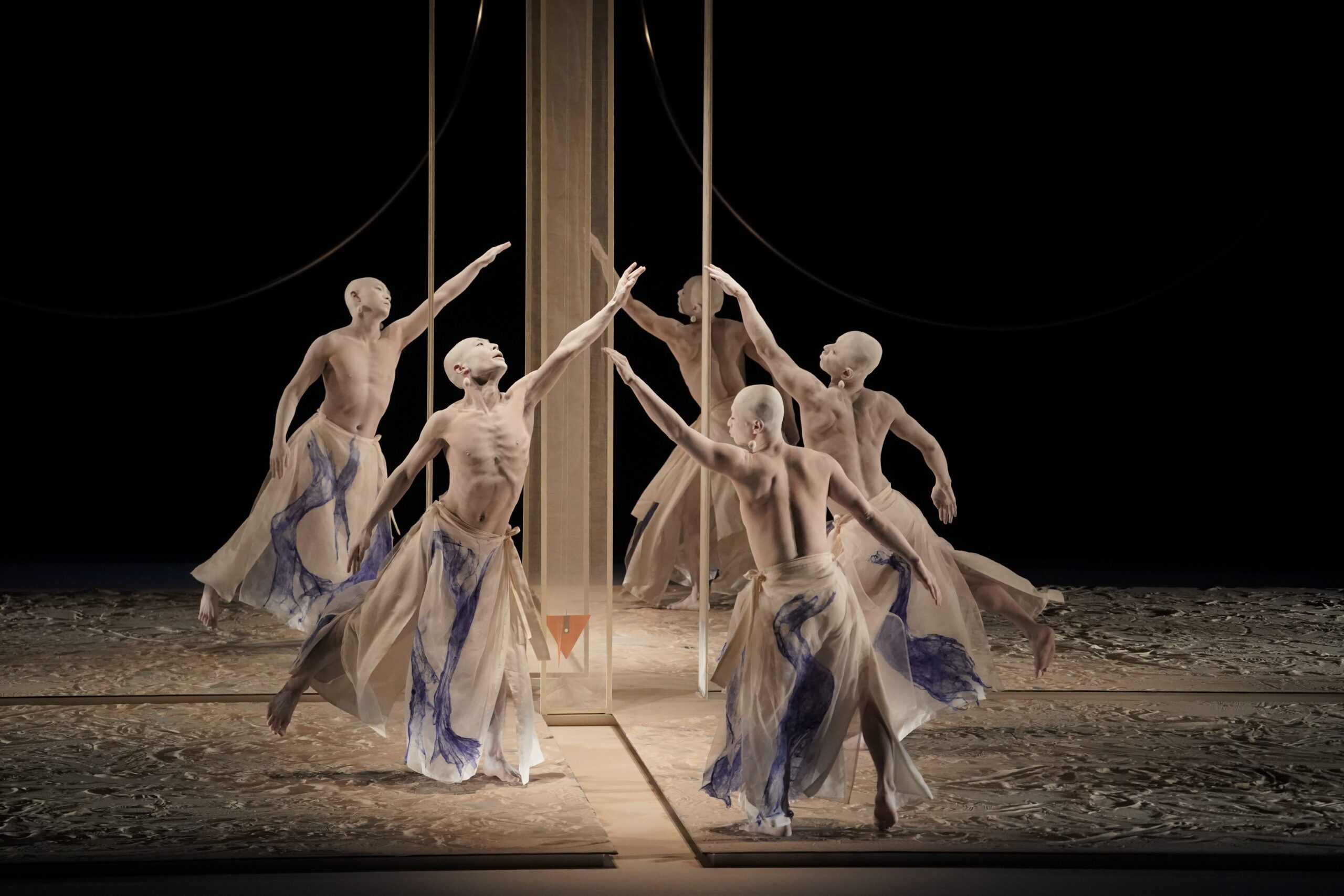 Five dancers with bald heads and flowing cream and blue skirts perform a synchronized, expressive dance around a vertical structure on a textured stage, reaching out with extended arms in dramatic poses under soft lighting.