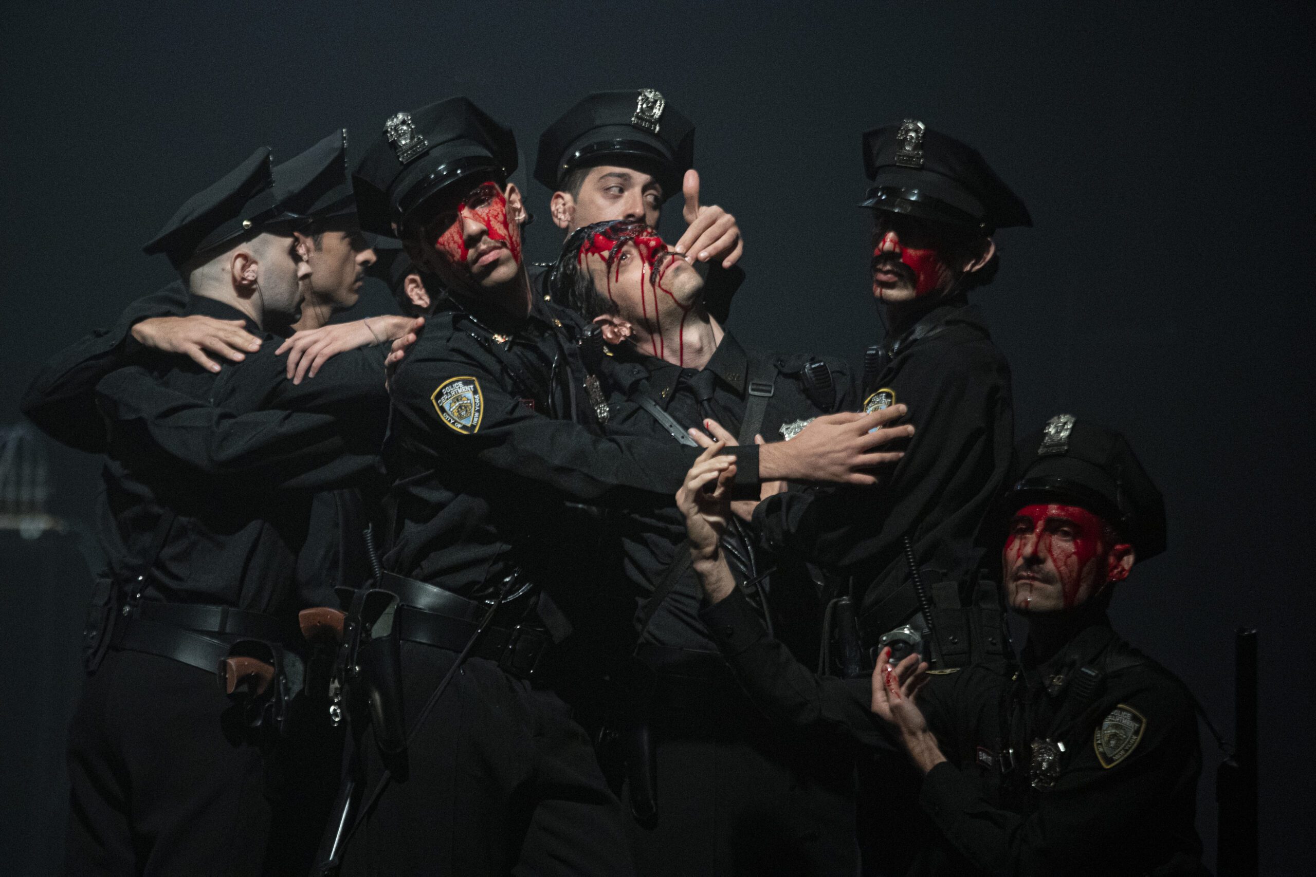 Six people in police uniforms stand close together, some with dramatic red face paint resembling blood. The lighting is dark and theatrical, evoking an intense, emotional scene.