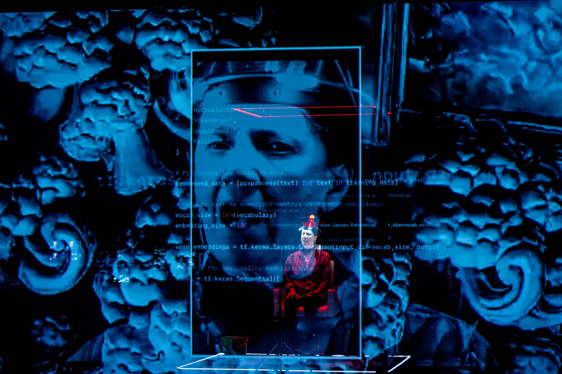 A person sits onstage framed by a glowing rectangle. Behind them is a blue-tinted image of a thoughtful face surrounded by brain imagery, overlaid with white programming code.