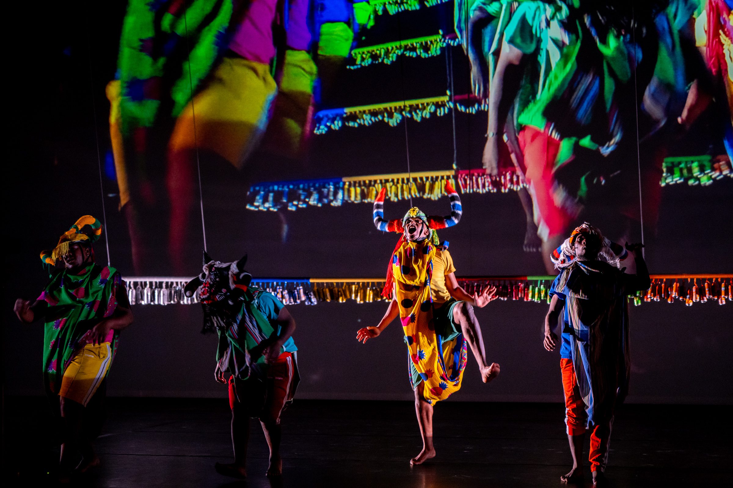 Four performers in colorful costumes and masks dance energetically on stage, with a vibrant projection of their movements displayed on the wall behind them. The scene is lively and theatrical.