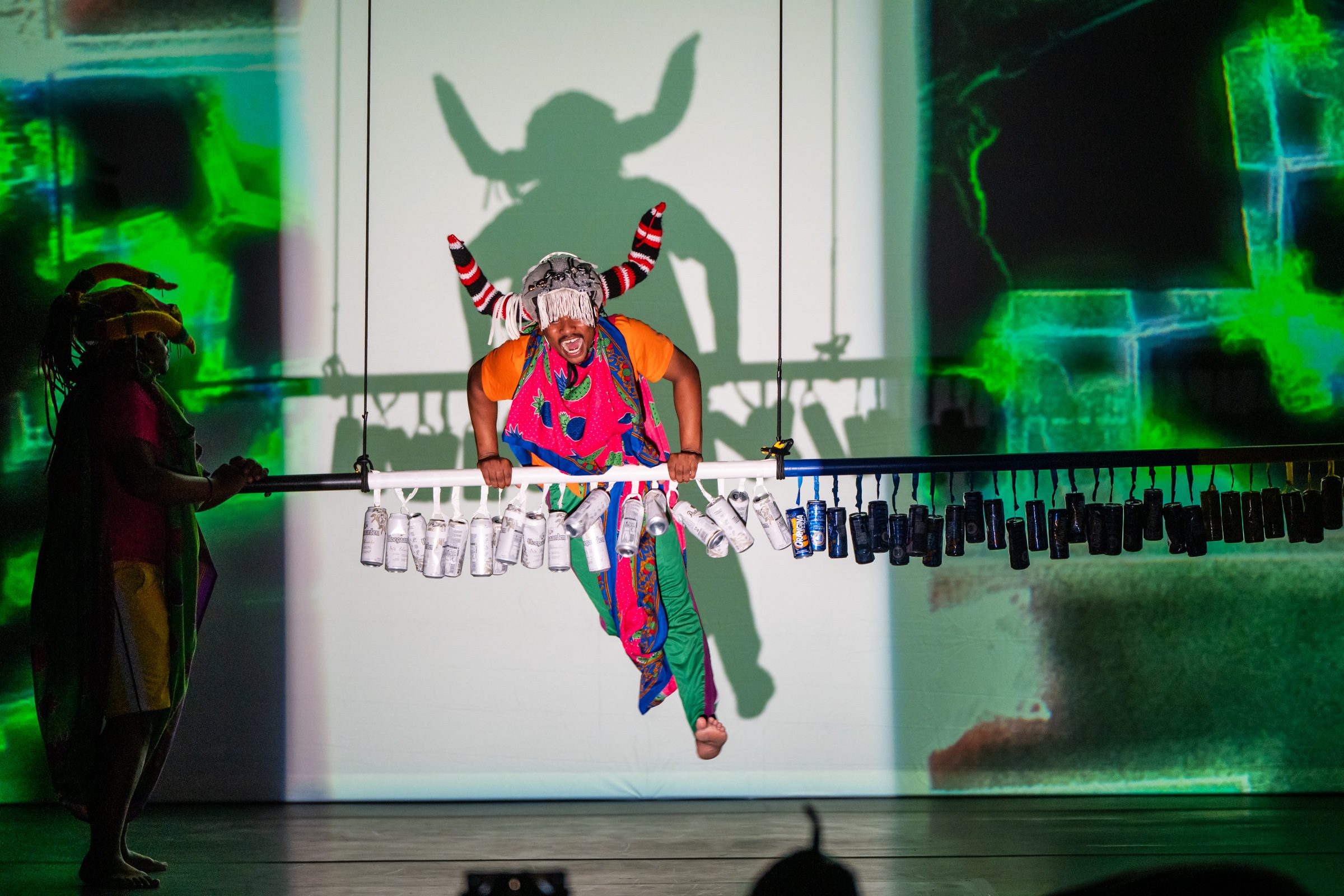 A performer in a colorful costume with horns and a mask hangs from a bar decorated with bottles, casting a dramatic shadow on a brightly lit, green and white abstract background.