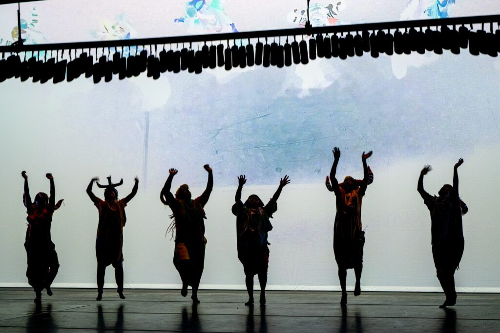 Six performers are silhouetted on stage, dancing with arms raised against a bright, abstract-lit background, under a row of hanging objects.