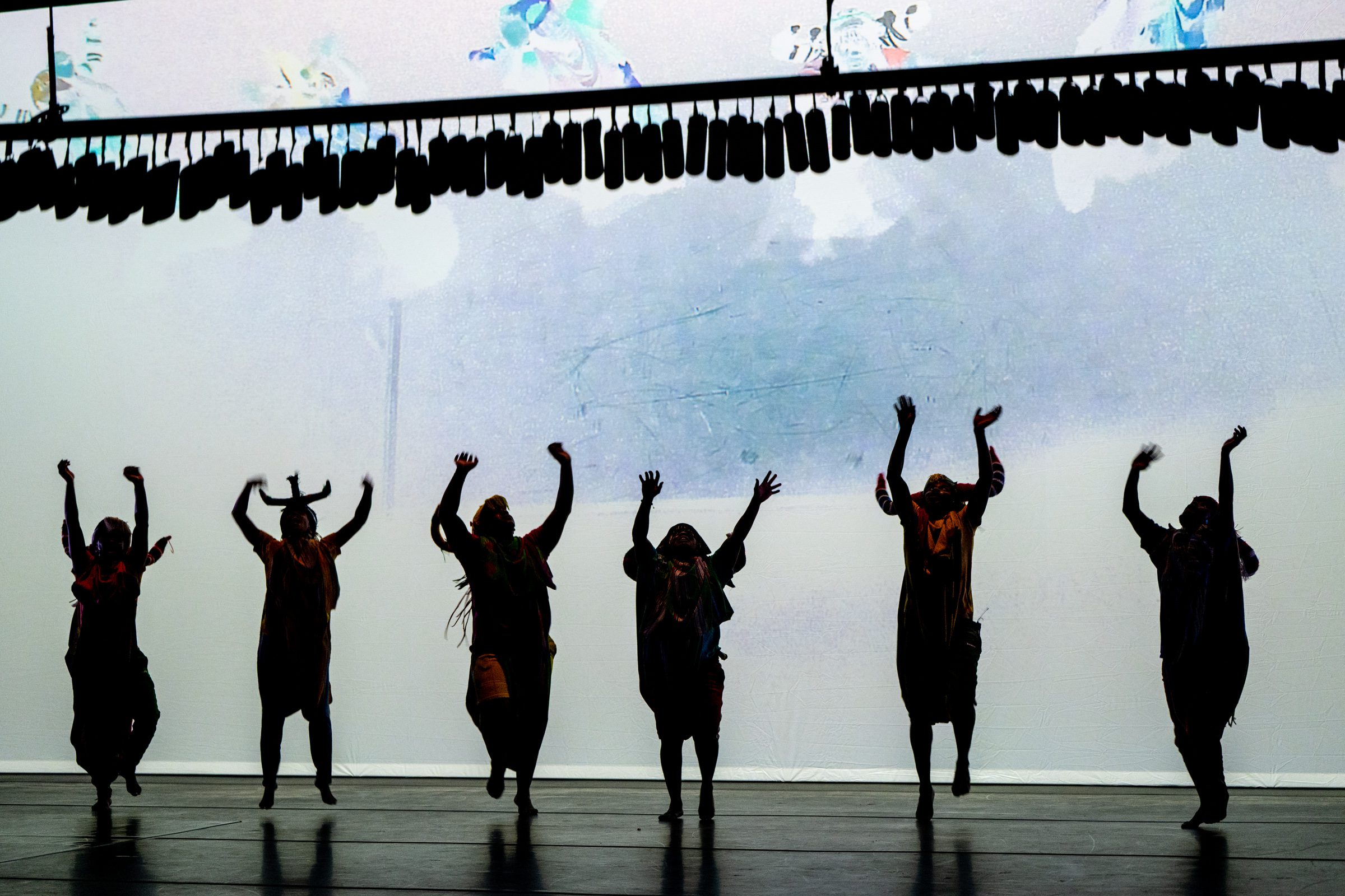 Six performers are silhouetted on stage, dancing with arms raised against a bright, abstract-lit background, under a row of hanging objects.