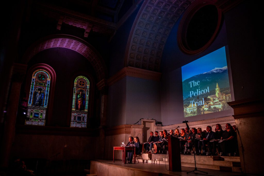 A person speaks at a table on stage in a grand hall with stained glass windows, while a group sits nearby and a large screen reads “The Pelicot Trial.”.