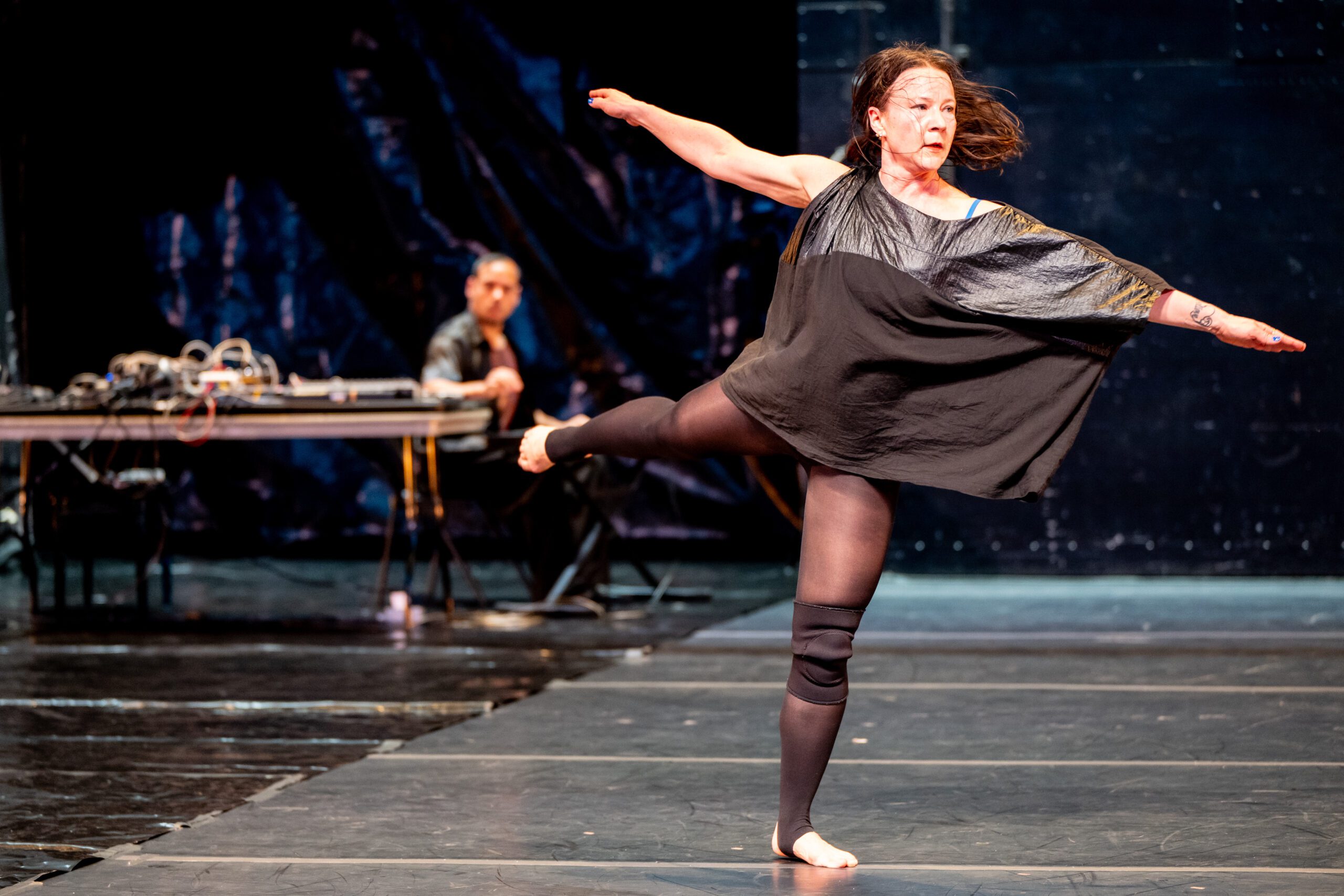 A woman in a black dress and tights performs a modern dance on stage, balancing on one leg with arms outstretched, while a person sits at a table with audio equipment in the background.