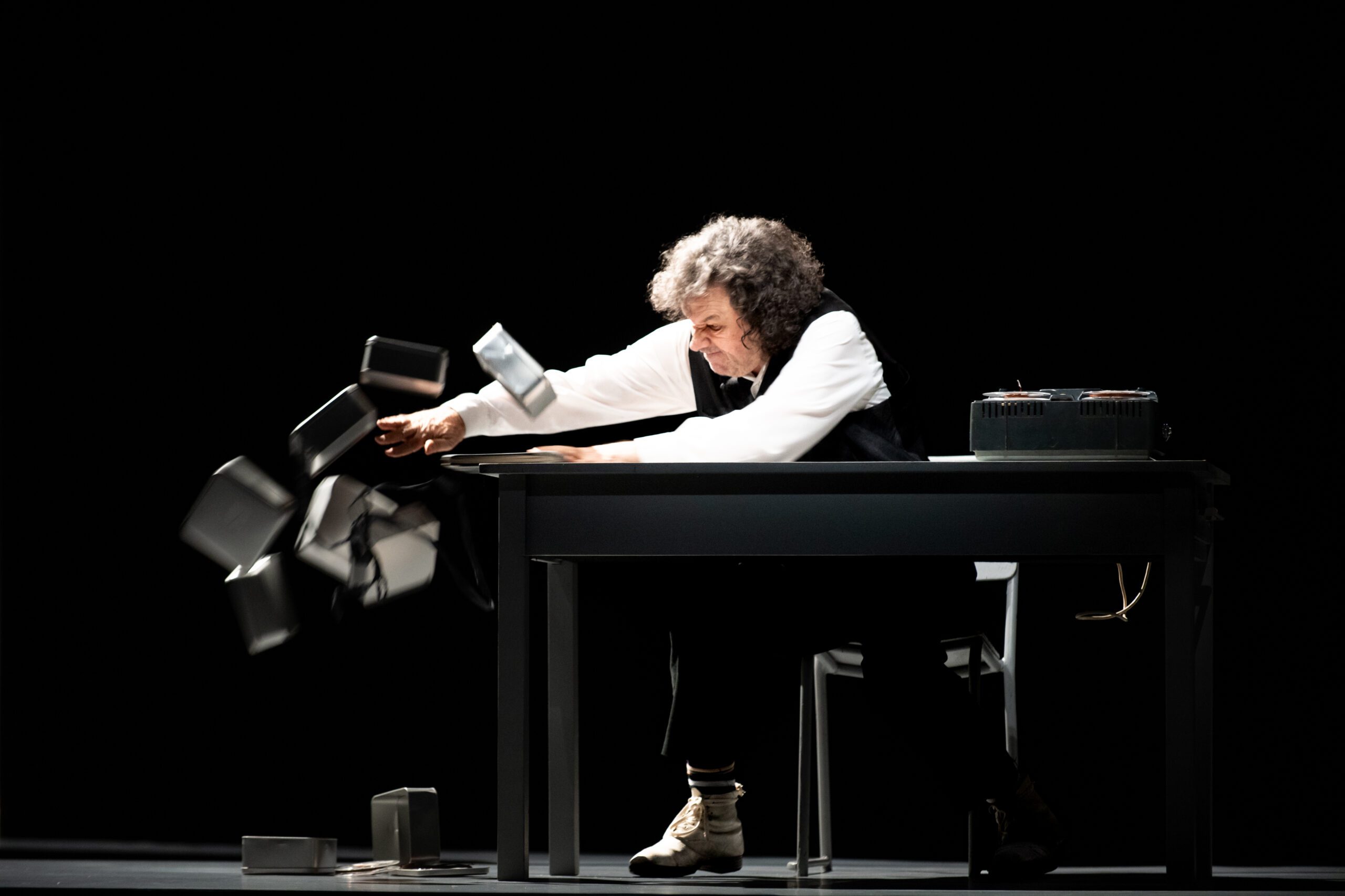 A person with curly hair sits at a table, knocking over several small rectangular objects with their arm. The scene is dramatically lit against a dark background.