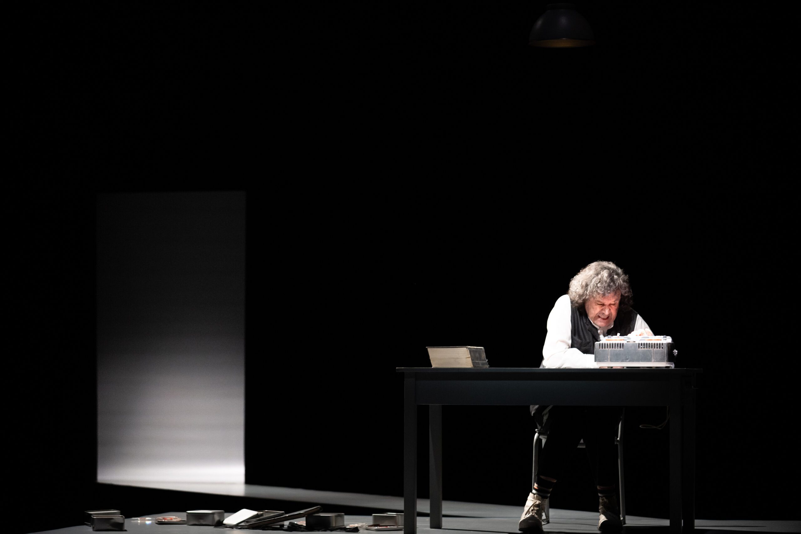 A person with curly hair sits alone at a dark table, leaning over audio equipment, with books and items scattered on the floor. A single overhead light illuminates the scene in an otherwise dark room.