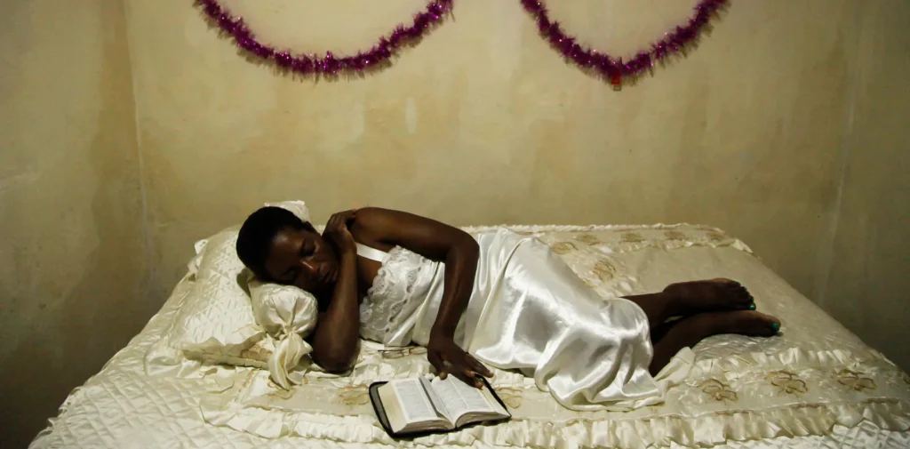 A woman in a white satin dress lies on a bed, resting her head on one arm with an open book in front of her. The wall behind her is decorated with purple garlands and bows.