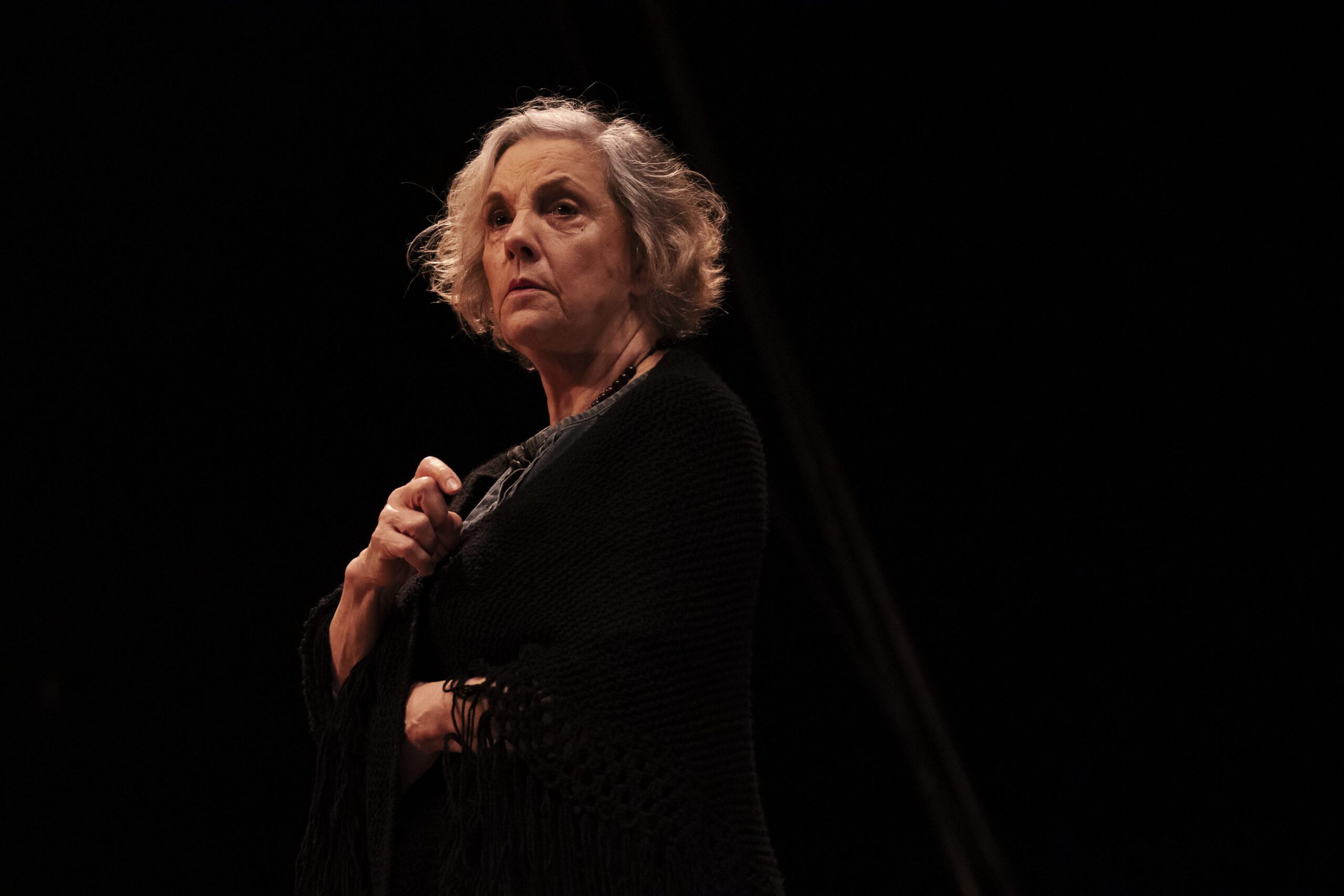 An older woman with short, wavy gray hair stands in dramatic lighting, wearing a dark shawl and looking intently off-camera against a black background.