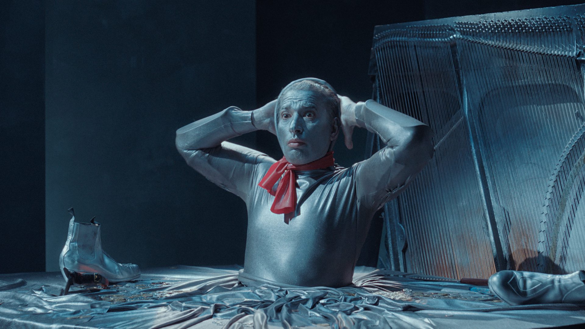 A person in silver body paint and clothing with a red scarf sits at a metallic table, holding their head with both hands. Silver boots are nearby, and a harp is in the background. The scene has a surreal, futuristic feel.