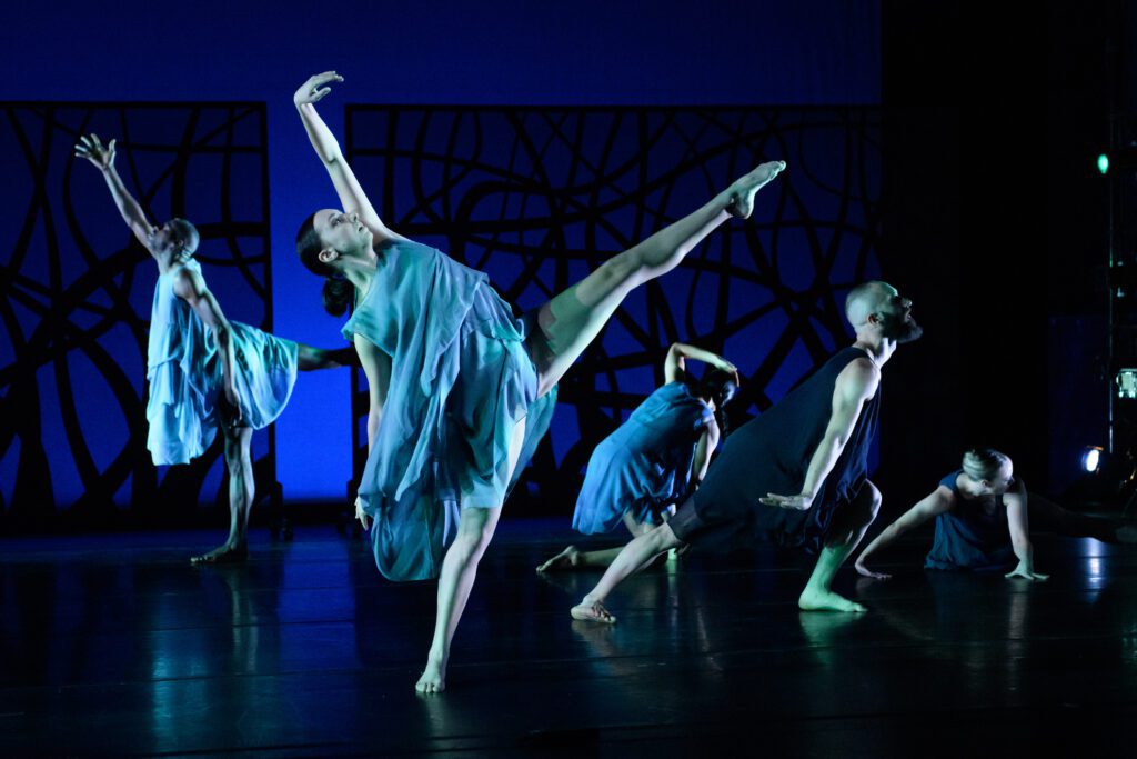 Five contemporary dancers in flowing blue costumes perform expressive movements on stage, with dramatic lighting and abstract black patterns in the background.