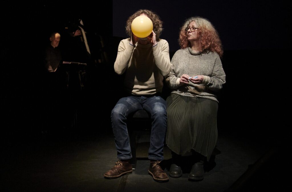 A person sits and inflates a yellow balloon, covering their face, while another person with curly hair and glasses sits beside them, watching. A musician is visible in the background.