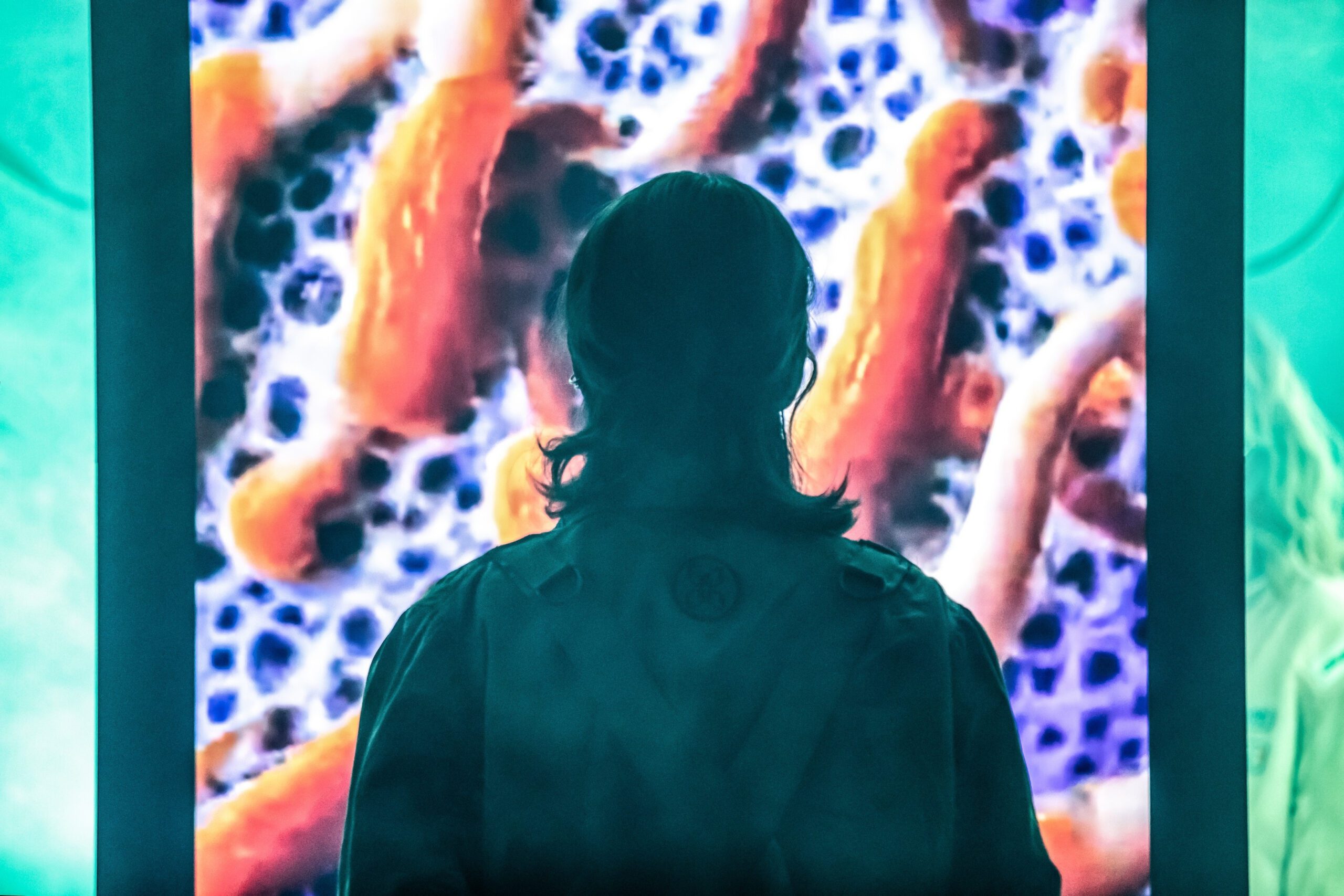 A person with long hair stands in front of a bright screen displaying a close-up image of orange and white microscopic organisms or cells.