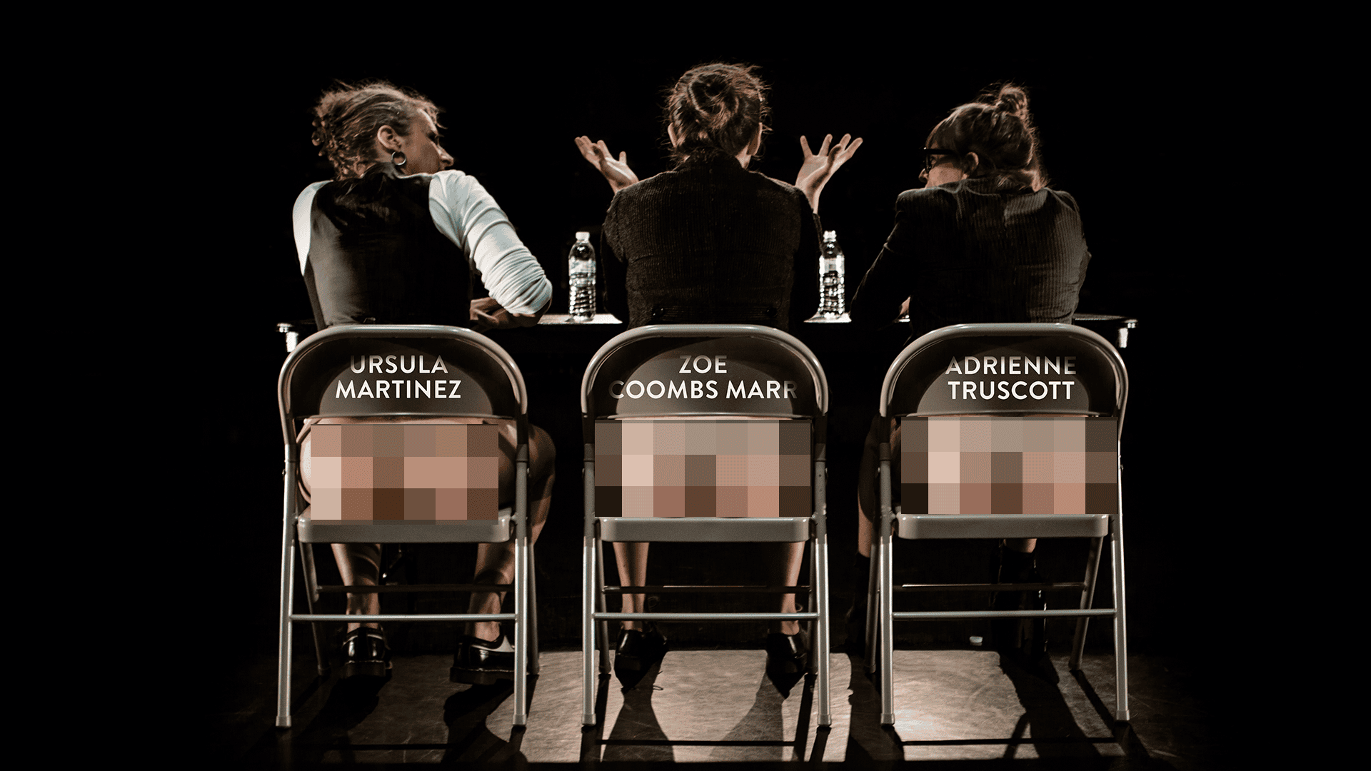 Three people sit on stage, backs to the camera, each on a chair labeled with their names. They are topless, but their lower backs are pixelated for modesty. Bottled water sits on their table.
