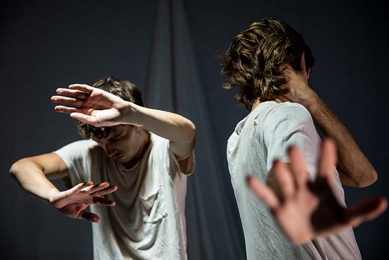 Two people in white shirts stand close together, raising their hands in front of their faces as if shielding themselves or dancing, against a dark, blurred background.