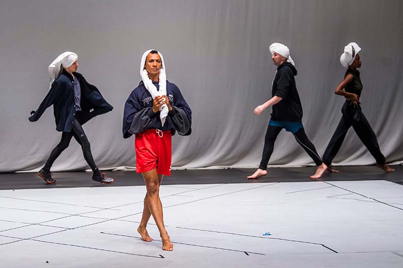 Four barefoot people wearing dark tops, light-colored towels on their heads, and shorts or leggings walk across a stage with a white backdrop and geometric lines on the floor. One person walks confidently in front.