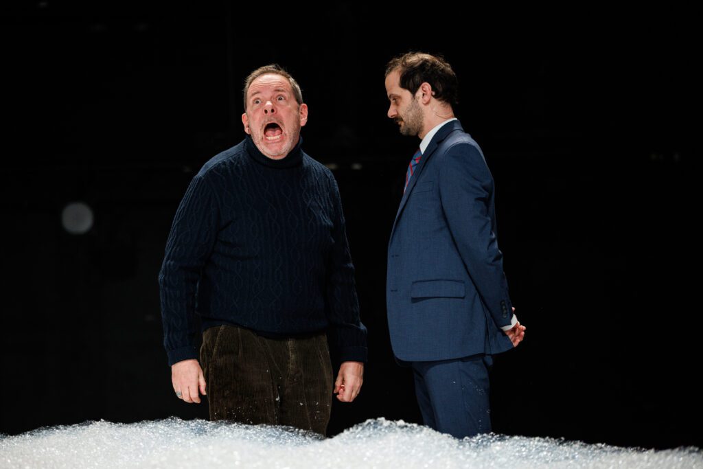 A man in a dark sweater shouts with his mouth wide open, while another man in a blue suit stands beside him, looking down with his hands behind his back.