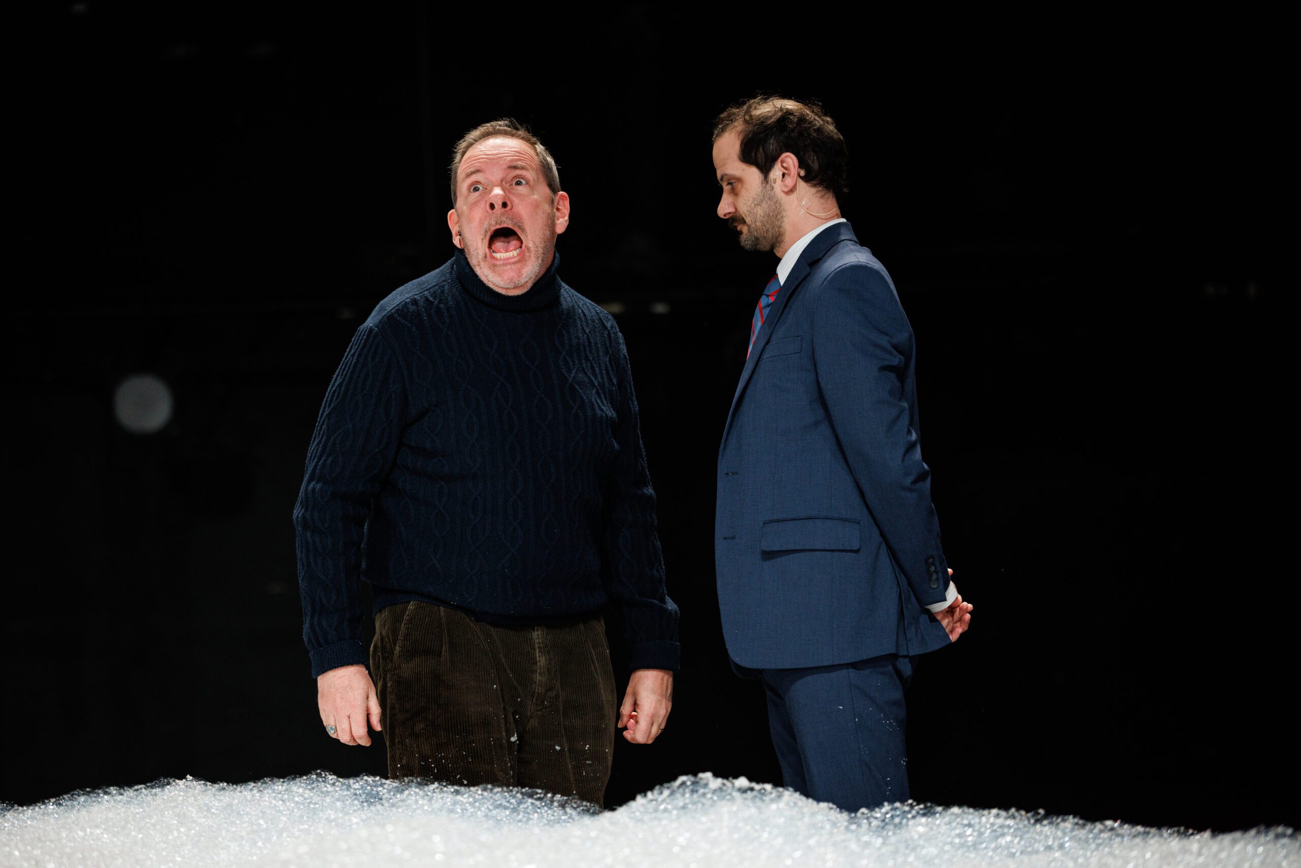 A man in a dark sweater shouts with his mouth wide open, while another man in a blue suit stands beside him, looking down with his hands behind his back.