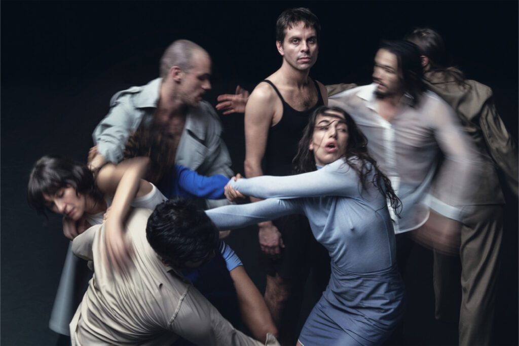 Six people, some blurred in motion, move around a man in black standing still at the center. The scene is dynamic and intense, with dramatic body language against a dark background.