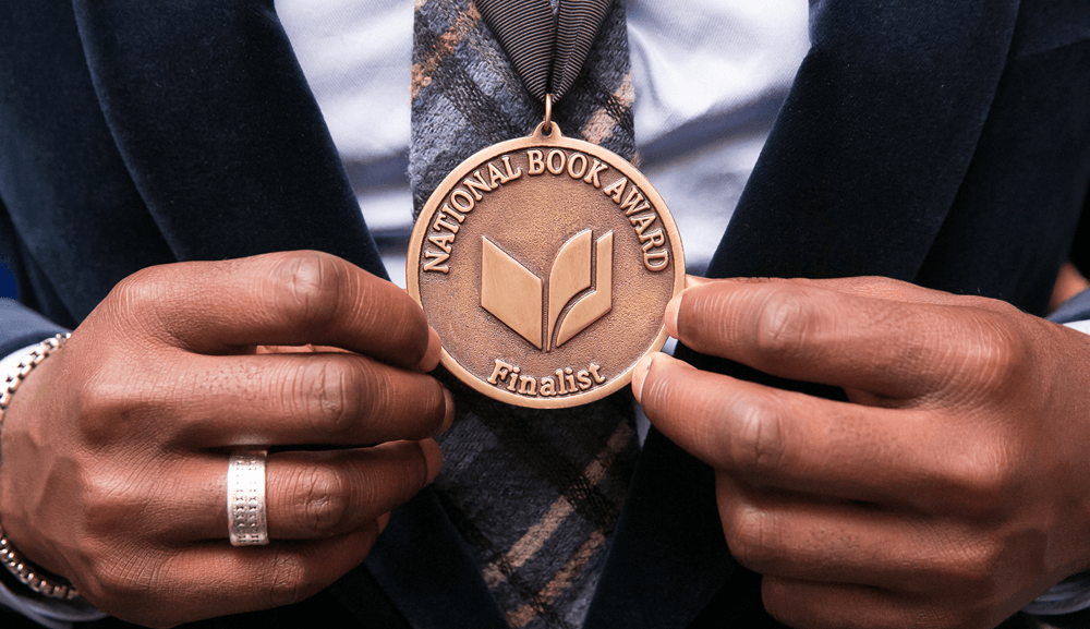 A person in a suit and plaid tie holds a large bronze medal reading “National Book Award Finalist” on a black ribbon.