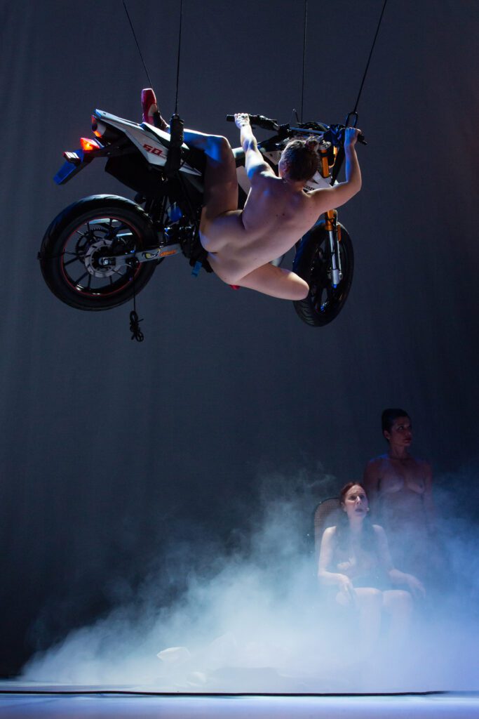 A nude person hangs upside down from a suspended motorcycle above a smoky stage, while two other people sit below, looking up.